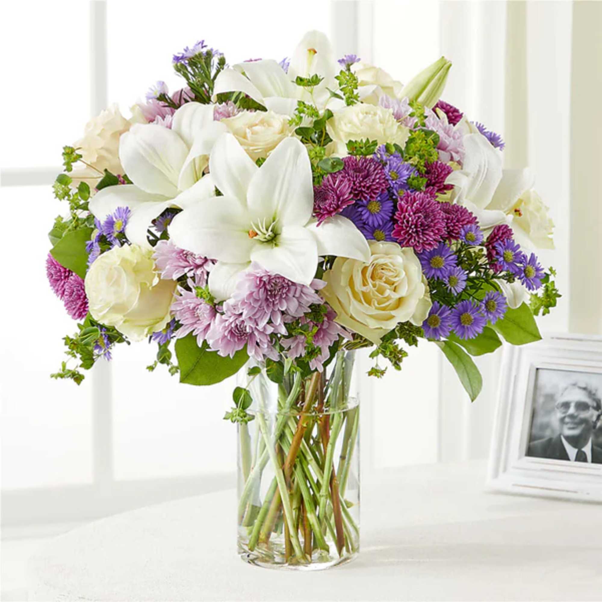 Bouquet of white lilies, cream roses, and purple daisies in a clear glass vase