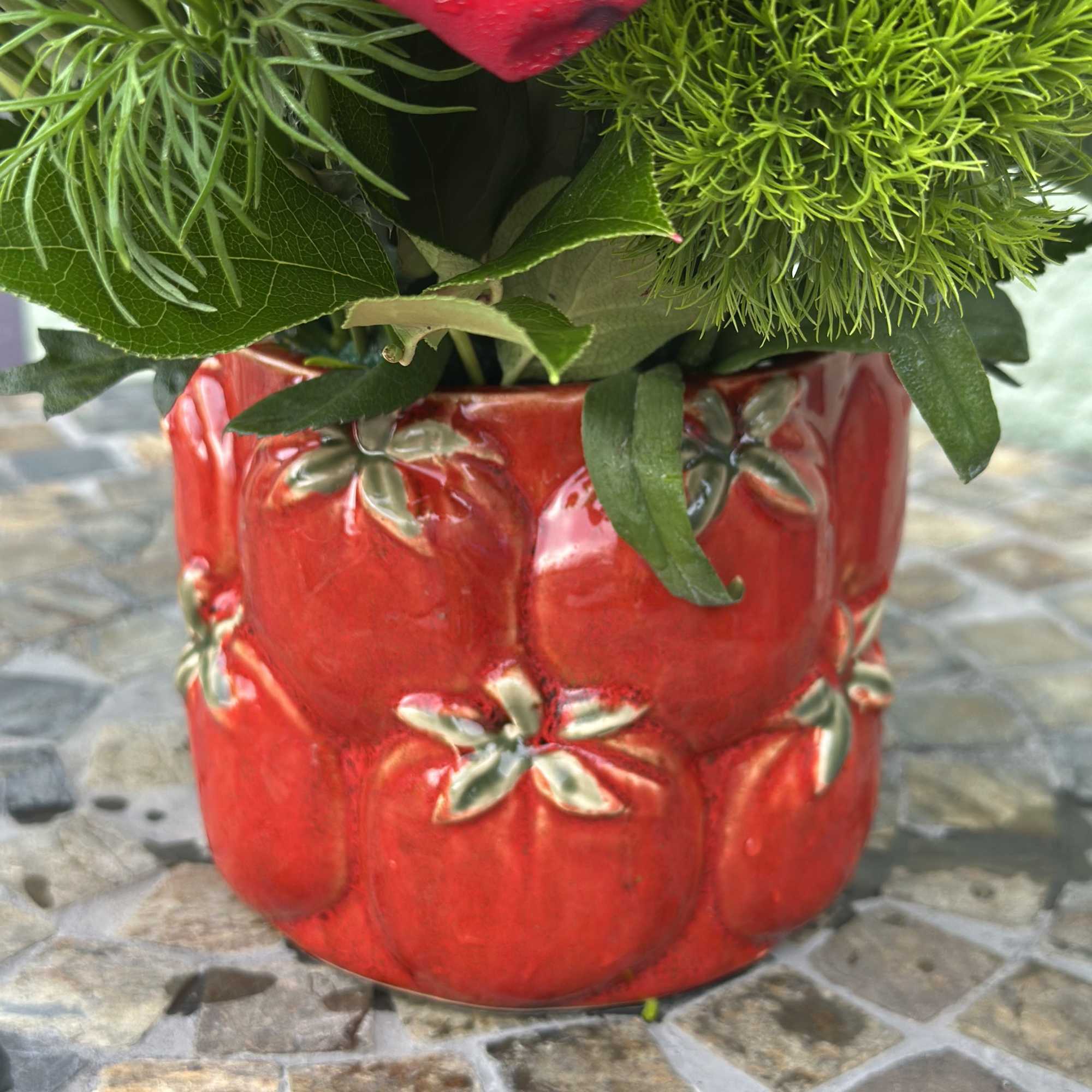 Short red ceramic pot with embossed tomatoes holding a red rose and bright green tufted blooms.