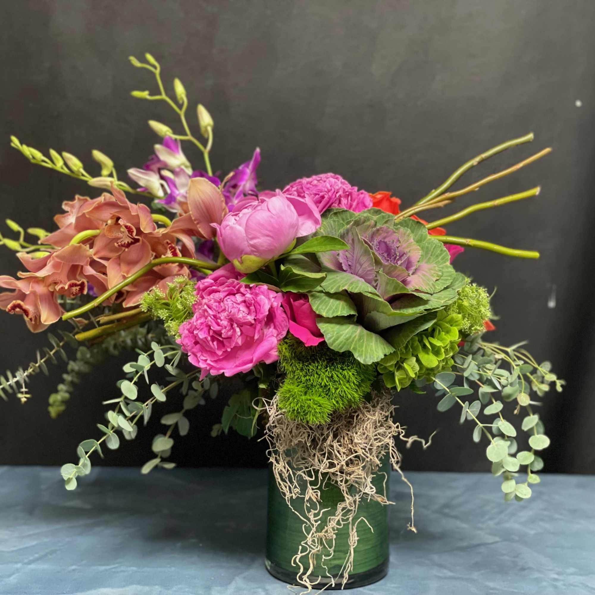Asymmetrical arrangement with pink peonies, peach orchids, and green accents in a glass cylinder vase
