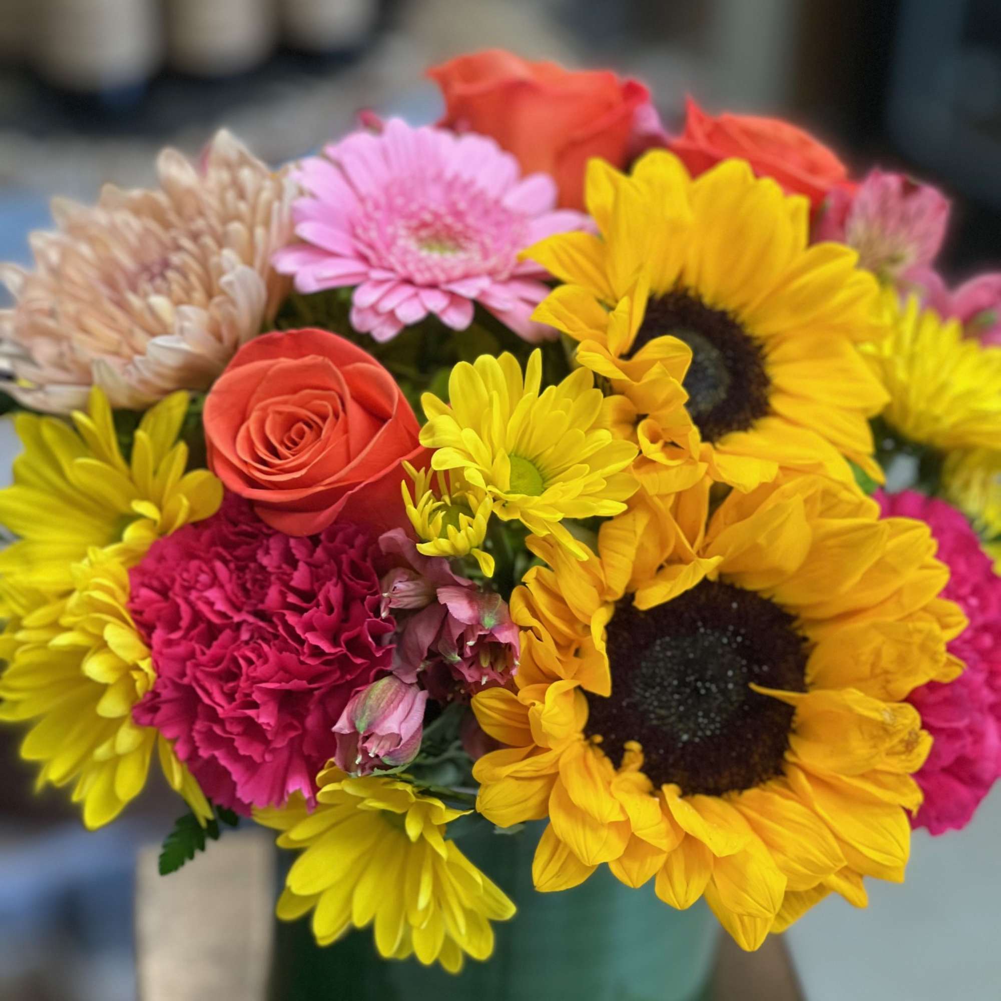 Bright bouquet of sunflowers, pink carnations, roses, and daisies in a clear glass vase.