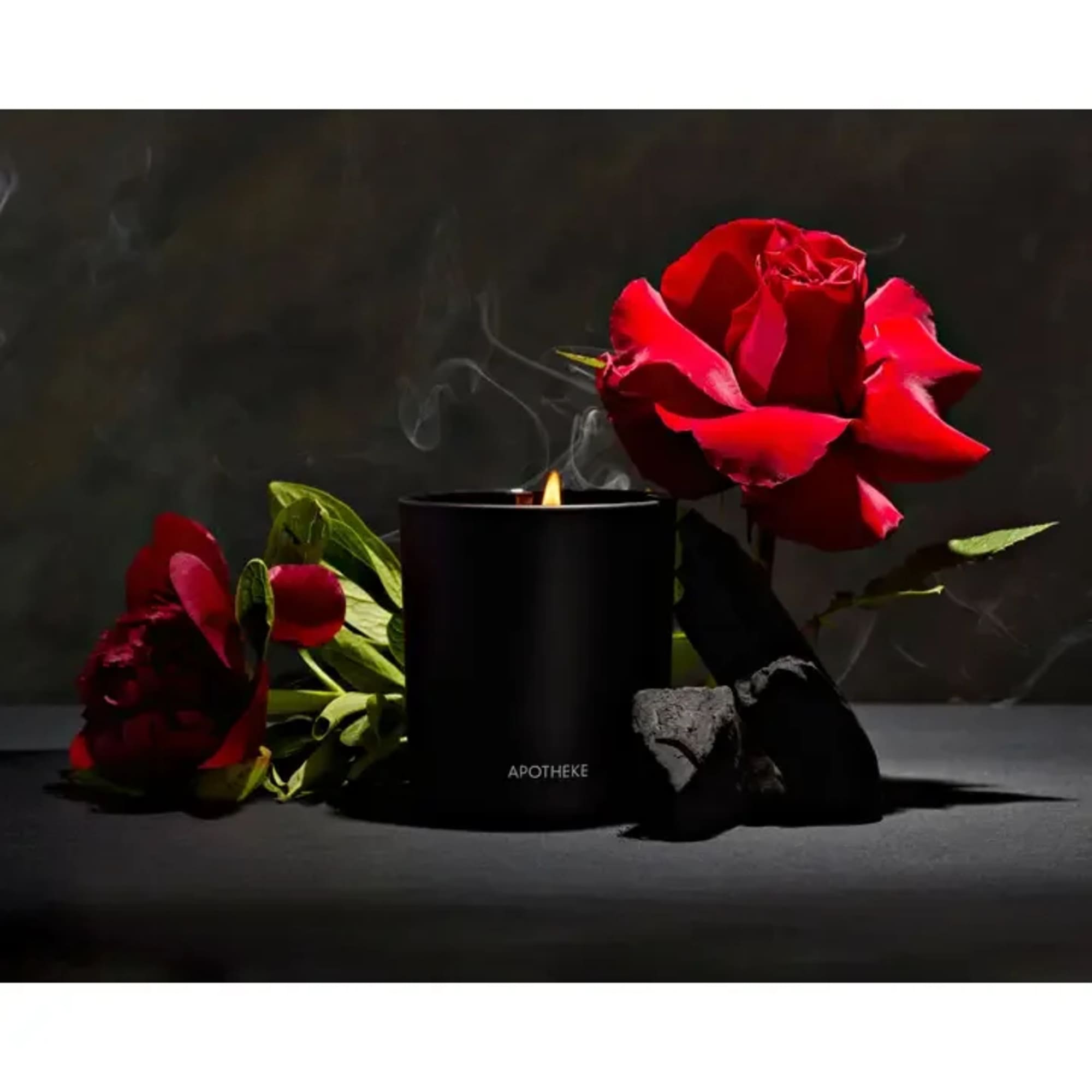 Black jar candle with flame beside red roses and dark stones on a dark surface