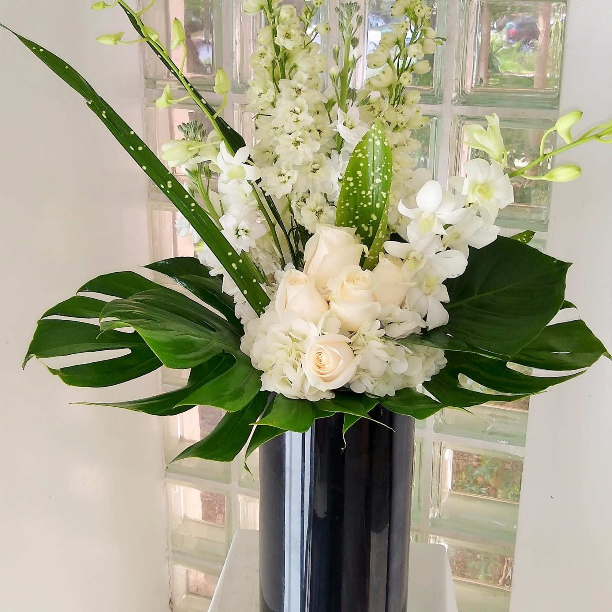 White floral arrangement in a tall black vase with large green leaves