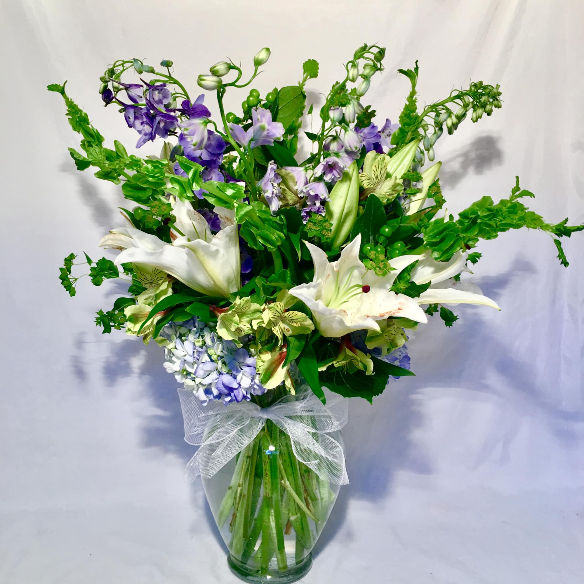 Tall arrangement of white lilies, green bells of Ireland, and purple flowers in a clear vase with a white bow