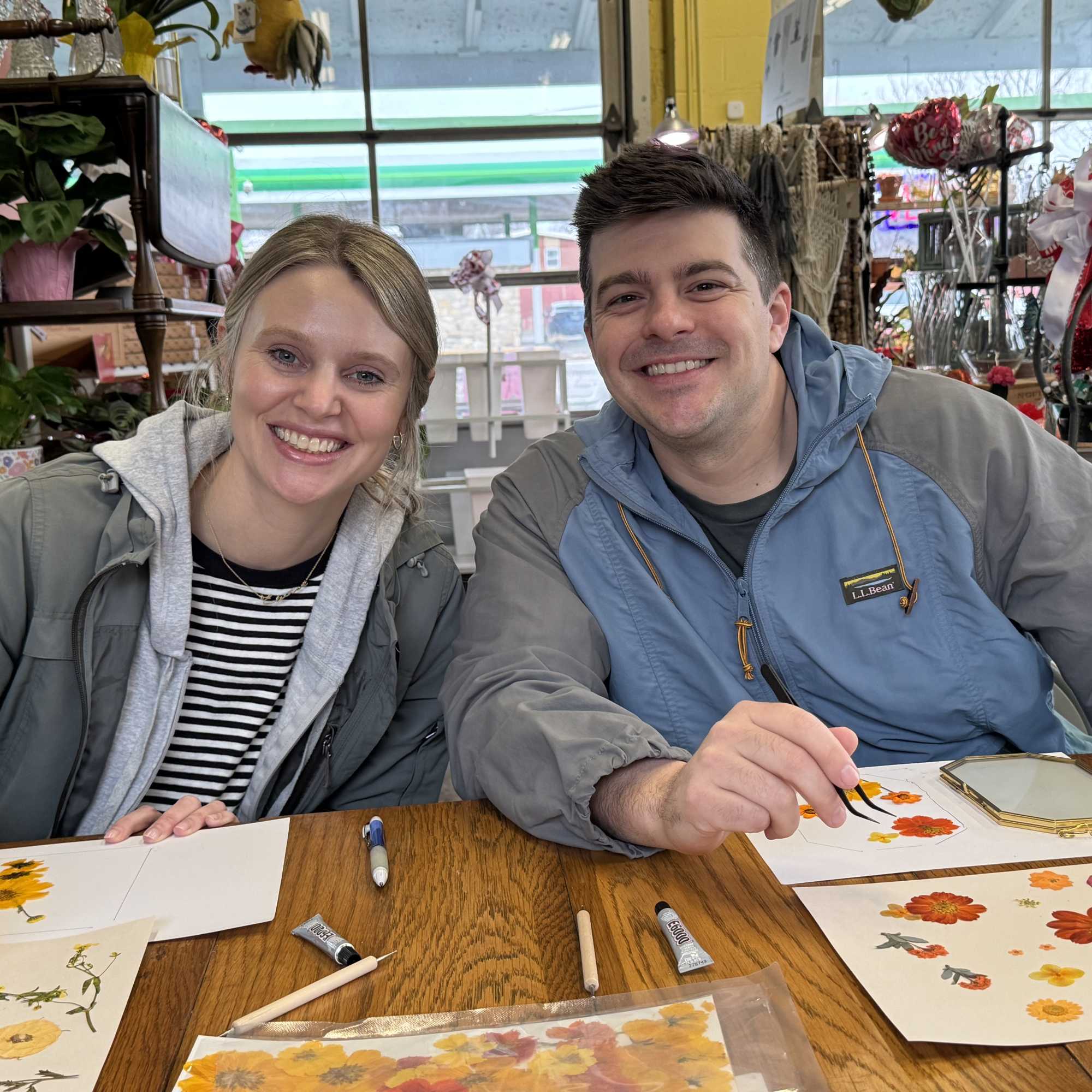 Two people sit at a table painting floral designs on paper.