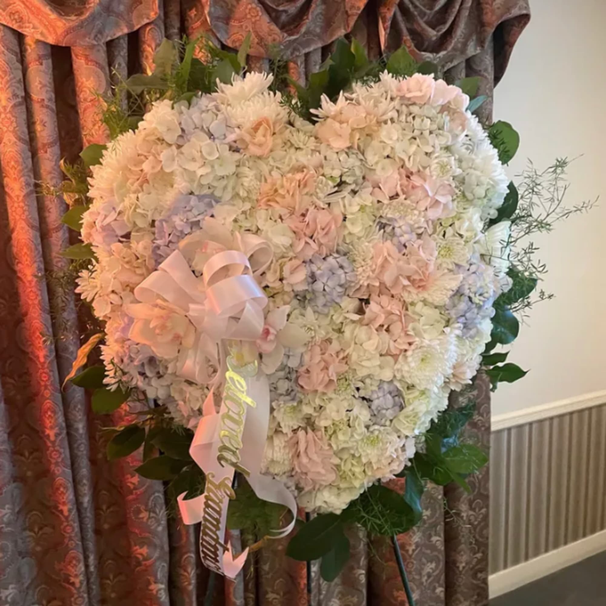 Large heart-shaped floral arrangement in pale pink, white, and lavender tones with a ribbon bow