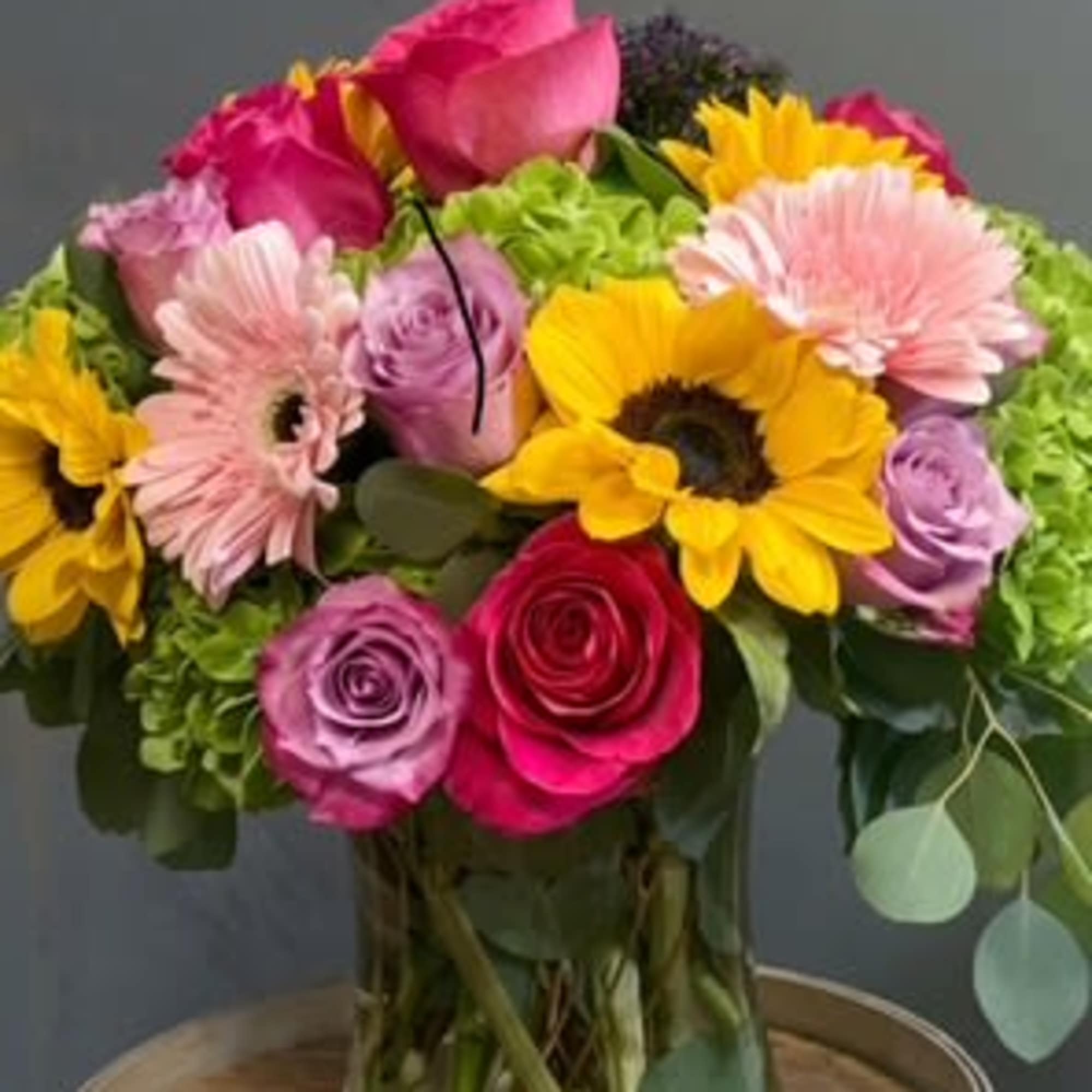 Bright mixed bouquet with sunflowers, pink roses, and gerbera daisies in a clear glass vase