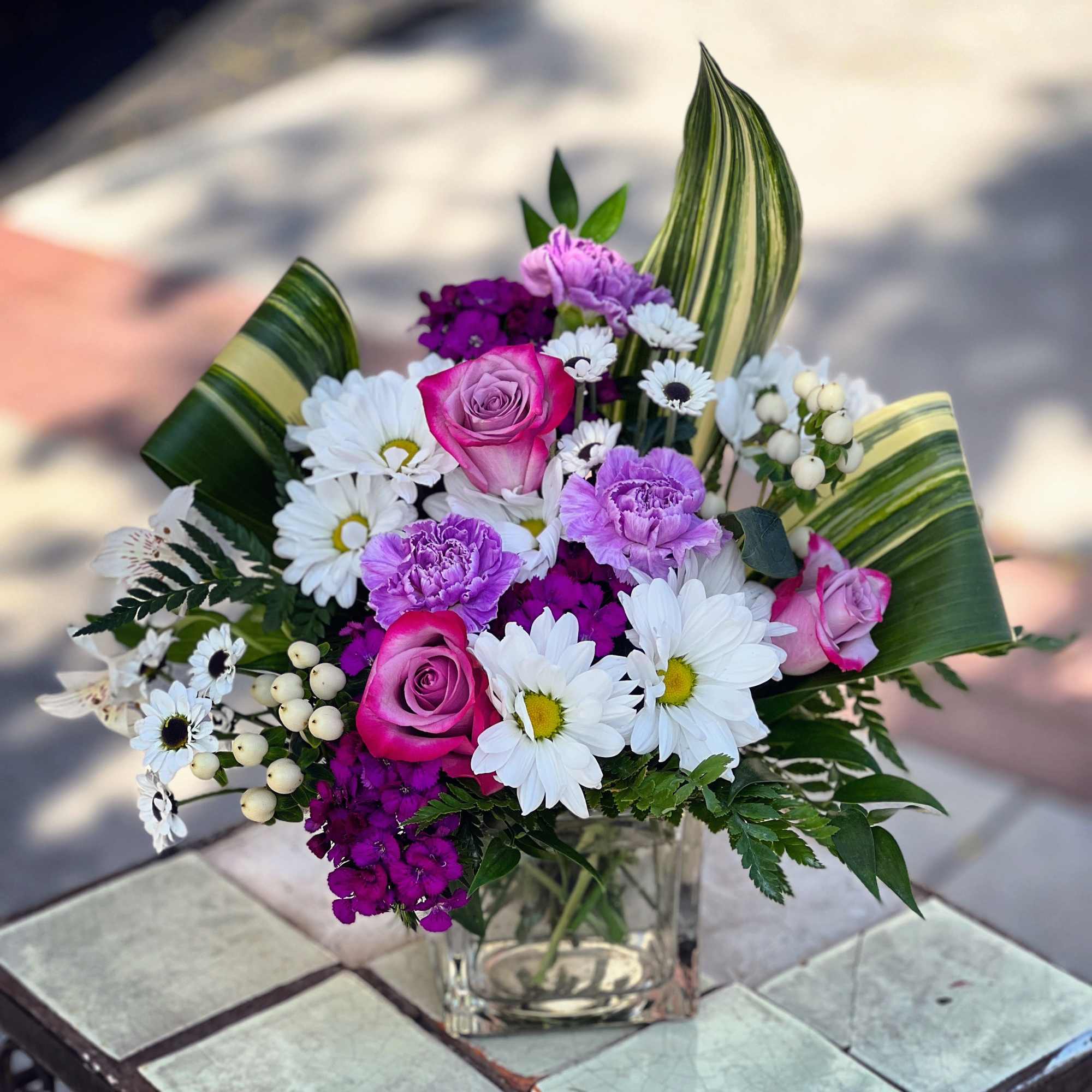 Pink roses and purple flowers in a clear glass vase with striped leaves