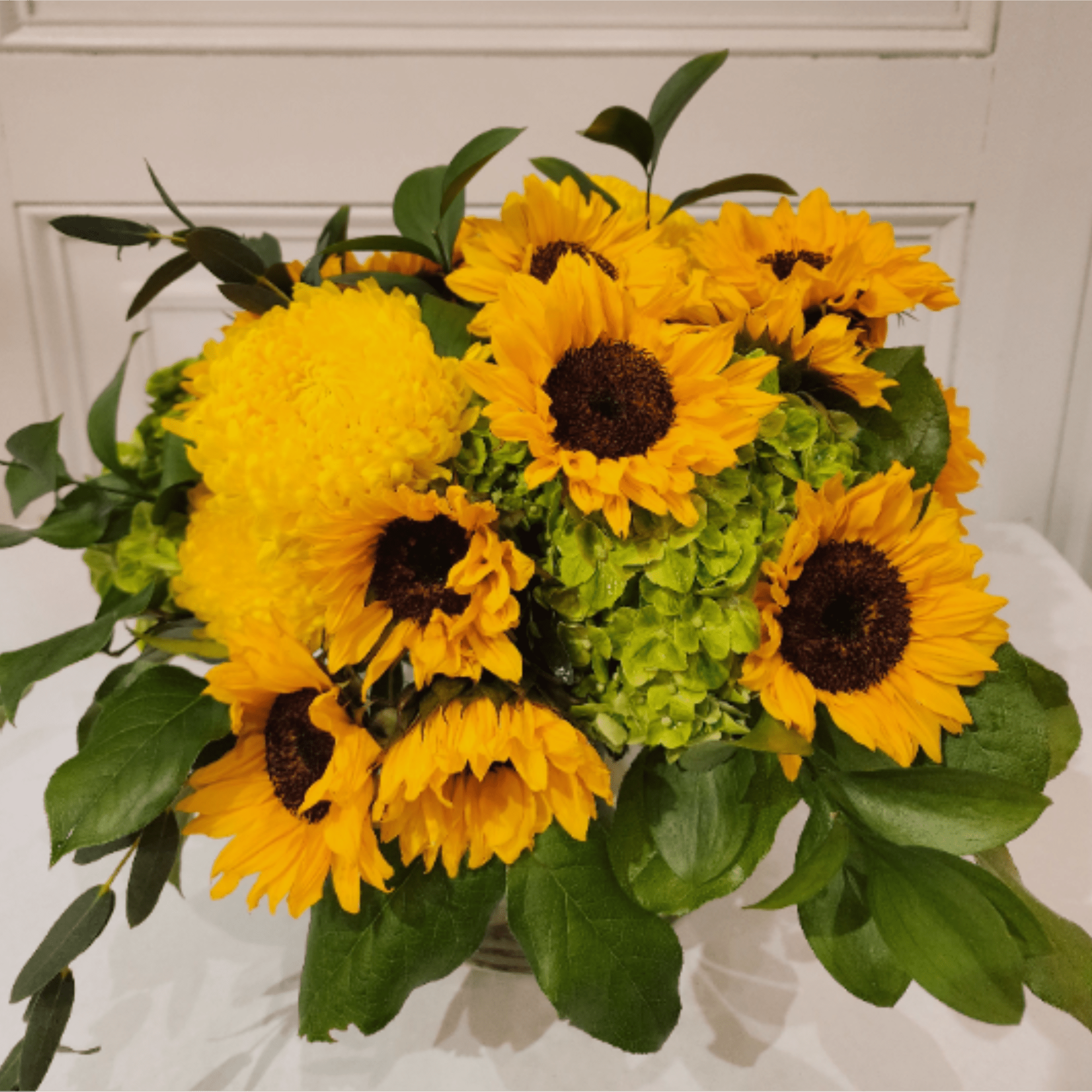 Low arrangement of yellow sunflowers and chrysanthemums with green hydrangeas in a clear glass vase