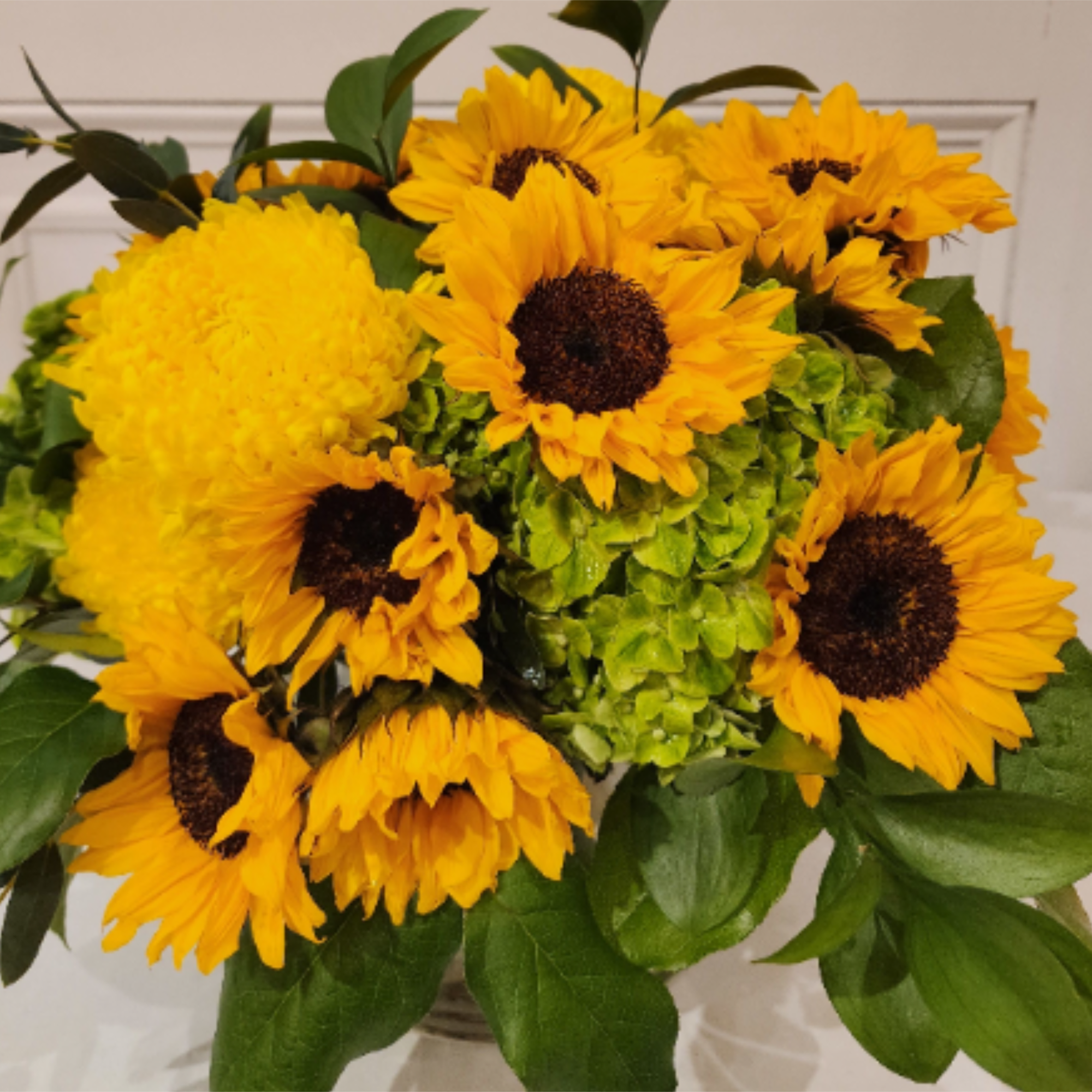Round arrangement of yellow sunflowers, yellow mum, and green hydrangeas in a clear glass vase