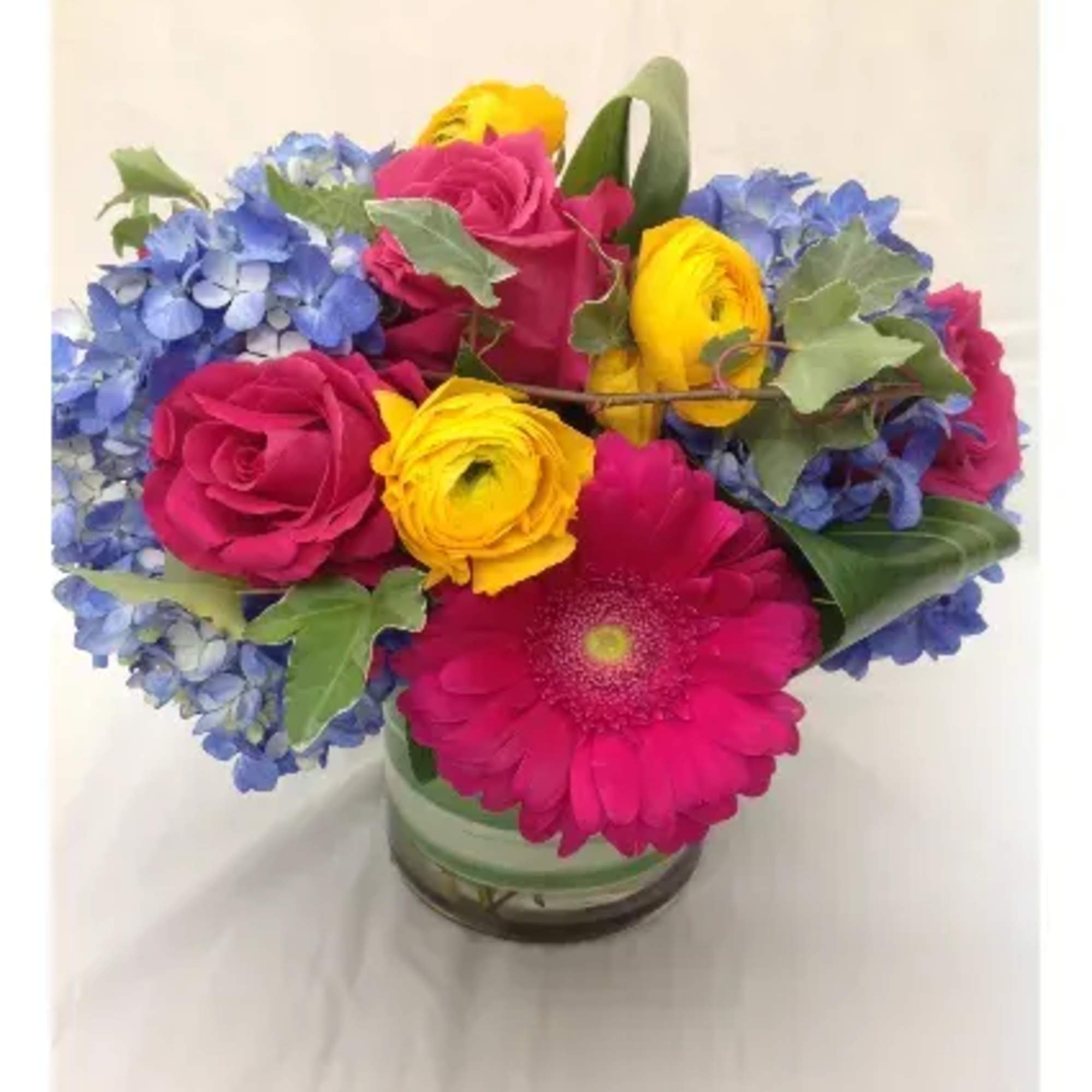 Bright arrangement of pink roses, yellow ranunculus, blue hydrangeas and a gerbera daisy in a glass vase