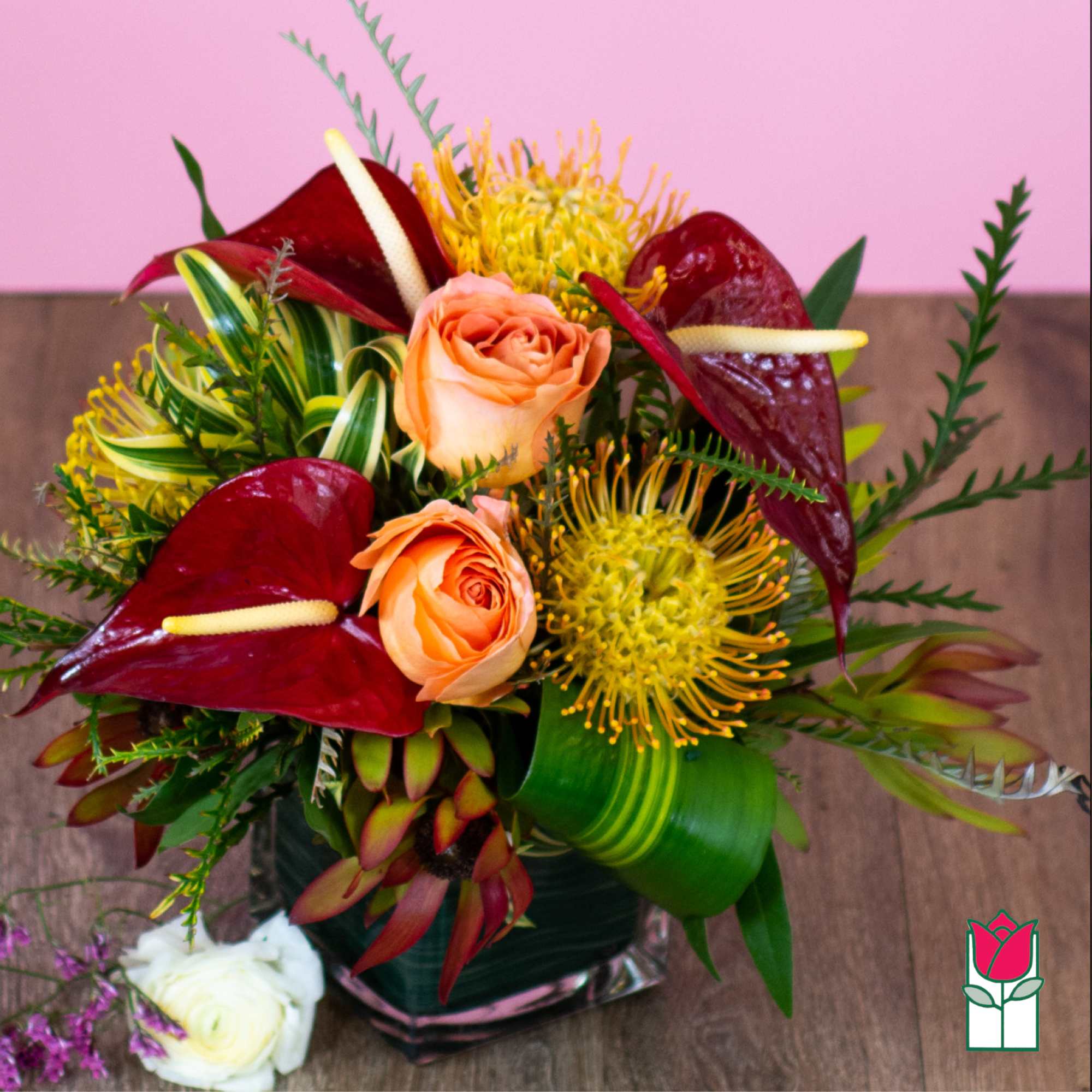 Tropical bouquet with peach roses, burgundy anthuriums, and yellow blooms in a glass vase