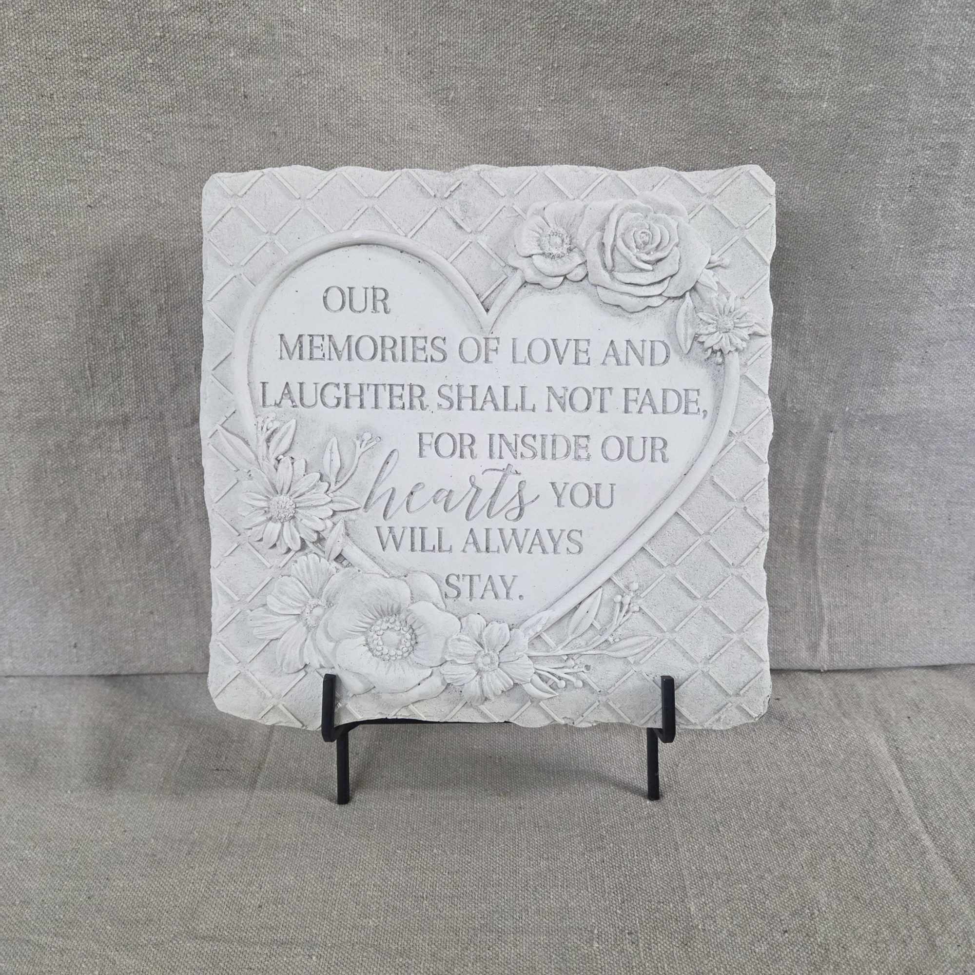 Square white memorial plaque with heart and sculpted flowers on a black metal stand.