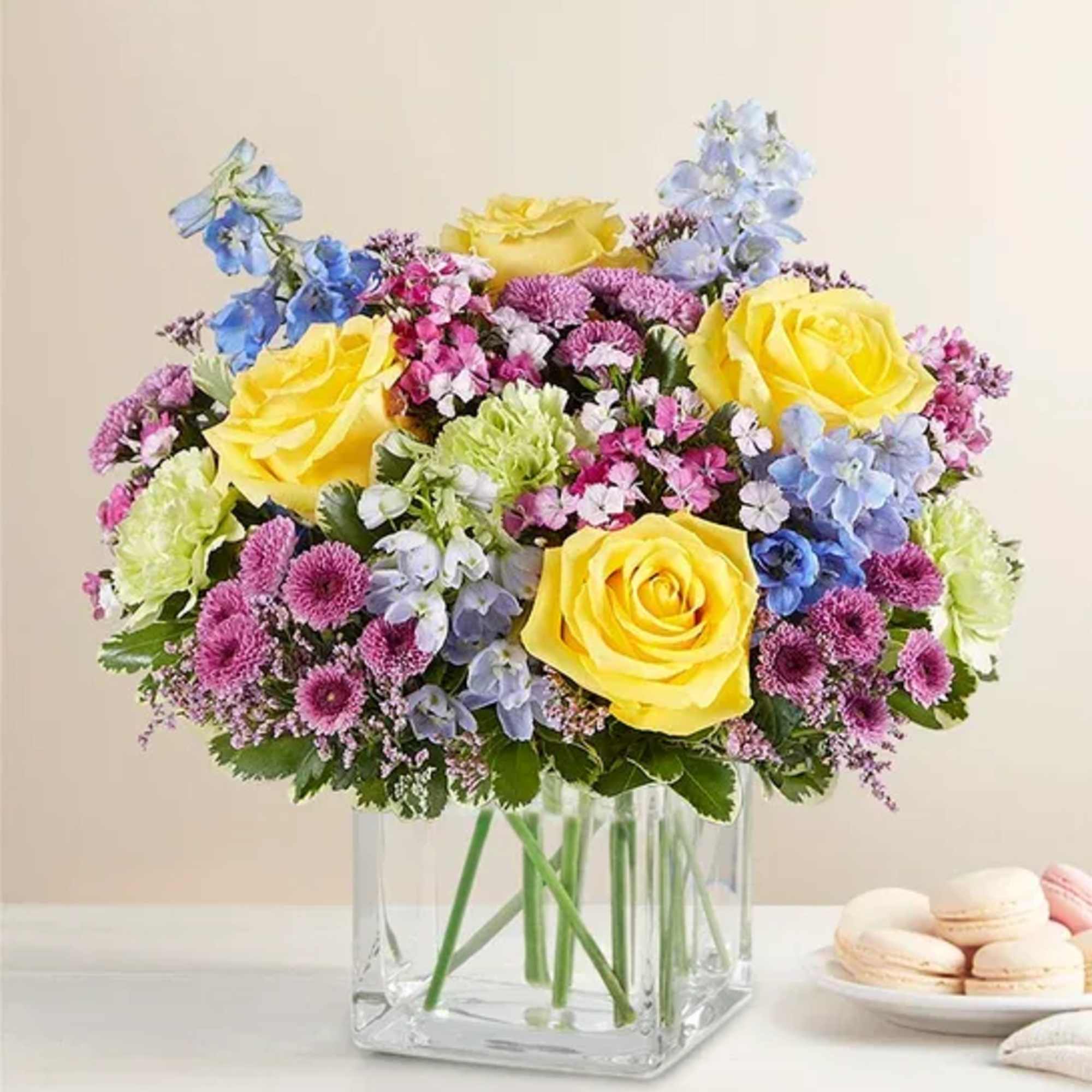 Mixed bouquet of yellow roses, blue flowers, and pink blooms in a clear glass vase