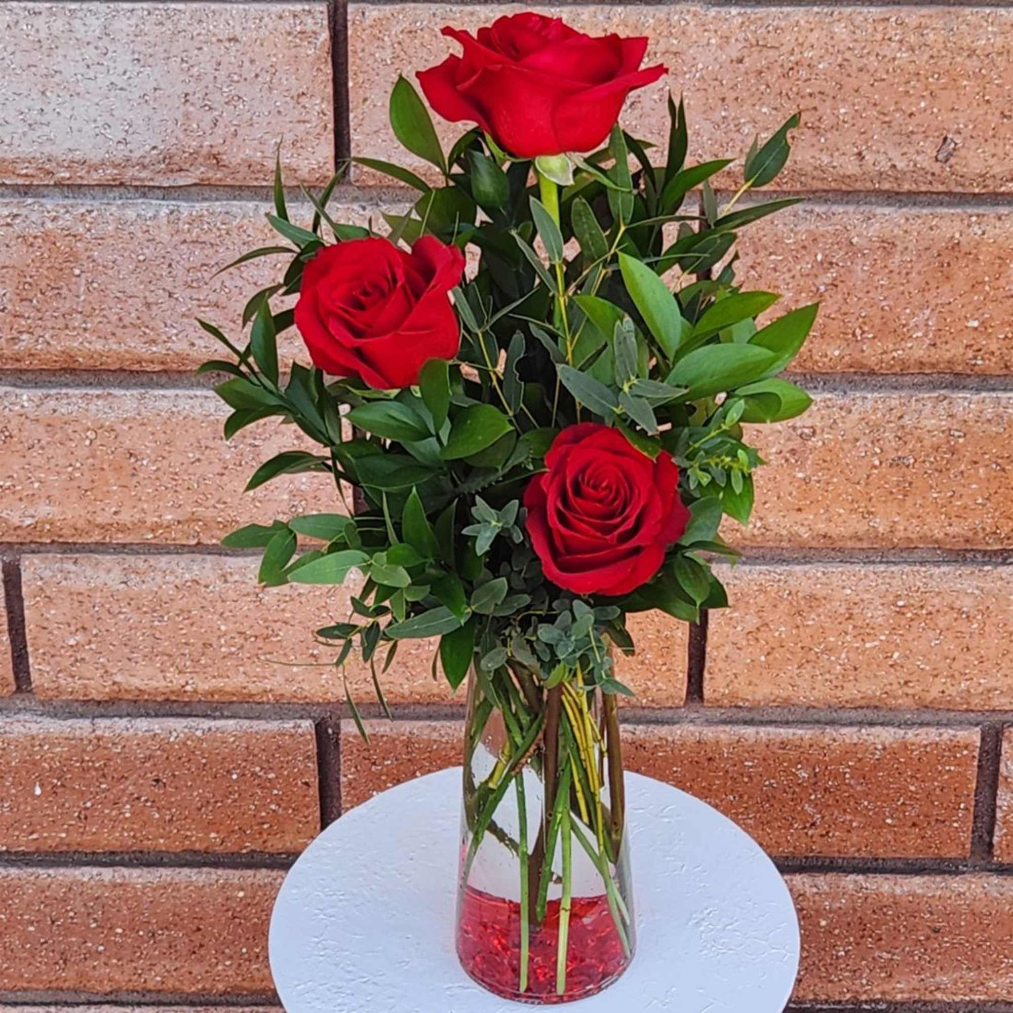 Three red roses in a clear glass vase with greenery