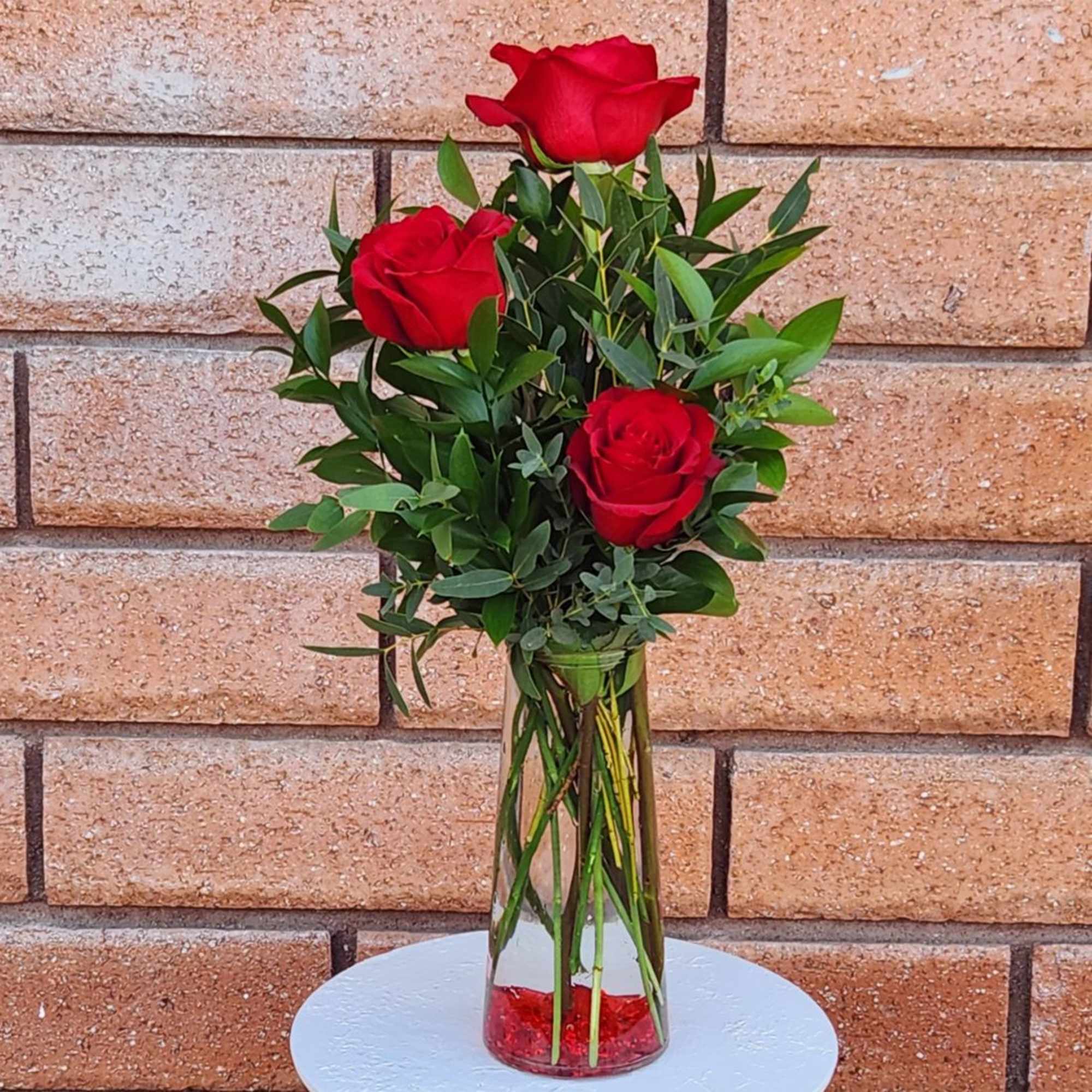 Three red roses in a clear glass vase with greenery