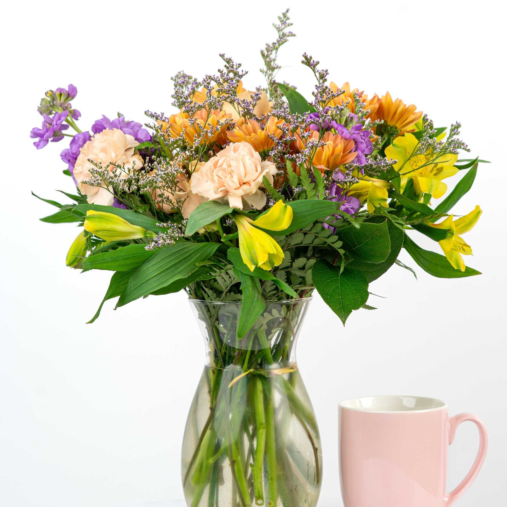 Mixed bouquet of peach, purple, and yellow flowers in a glass vase beside a pink mug