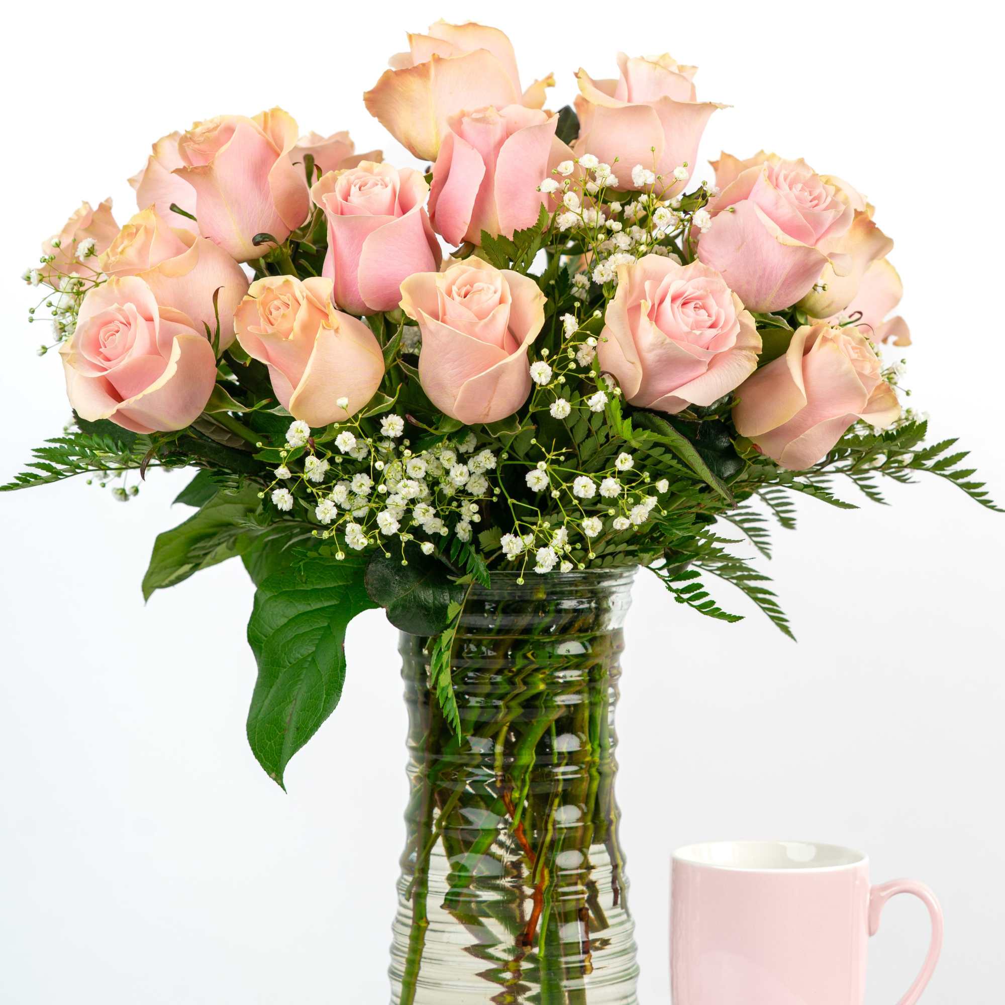 Tall glass vase of light pink roses with white filler flowers beside a pink mug