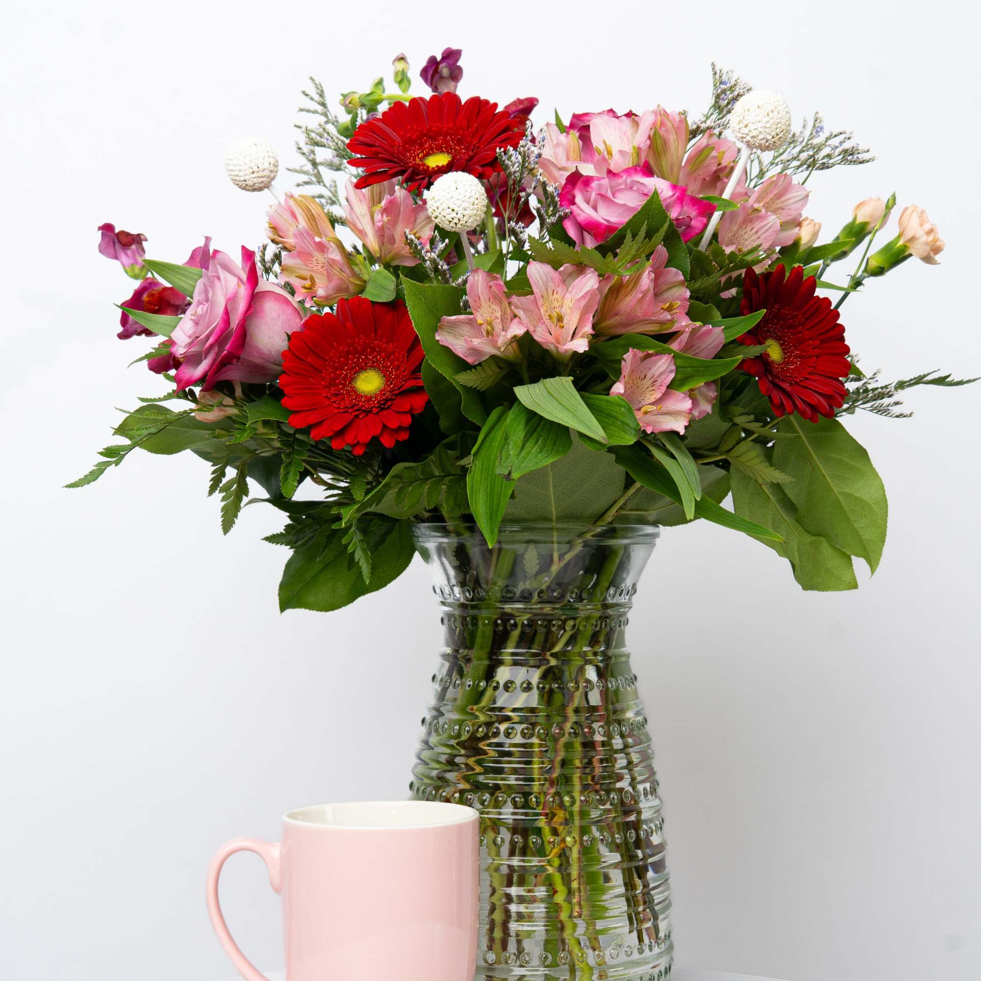 Pink and red mixed bouquet in a glass vase with a pink mug in front
