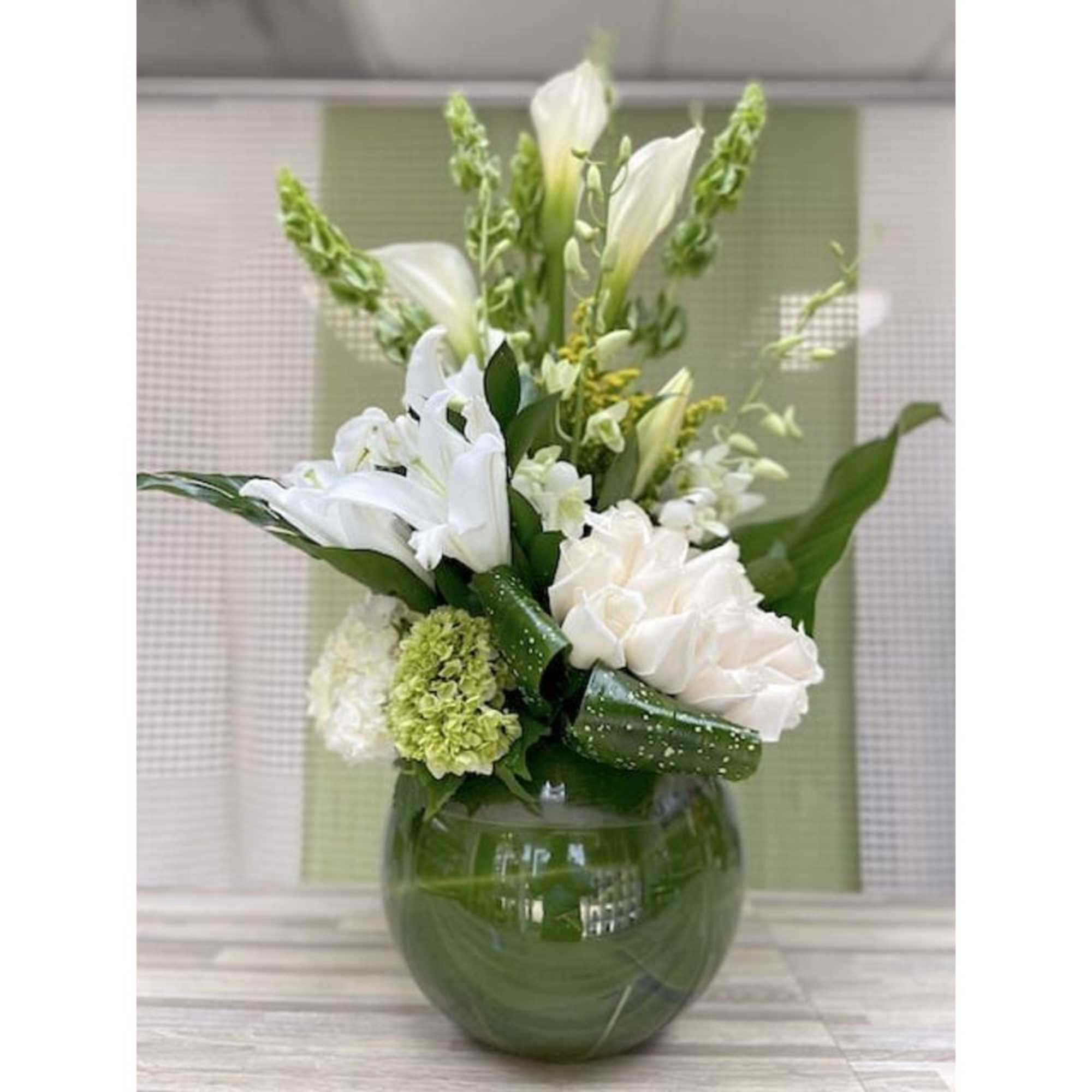 Tall arrangement of white lilies and roses with green accents in a round green glass vase