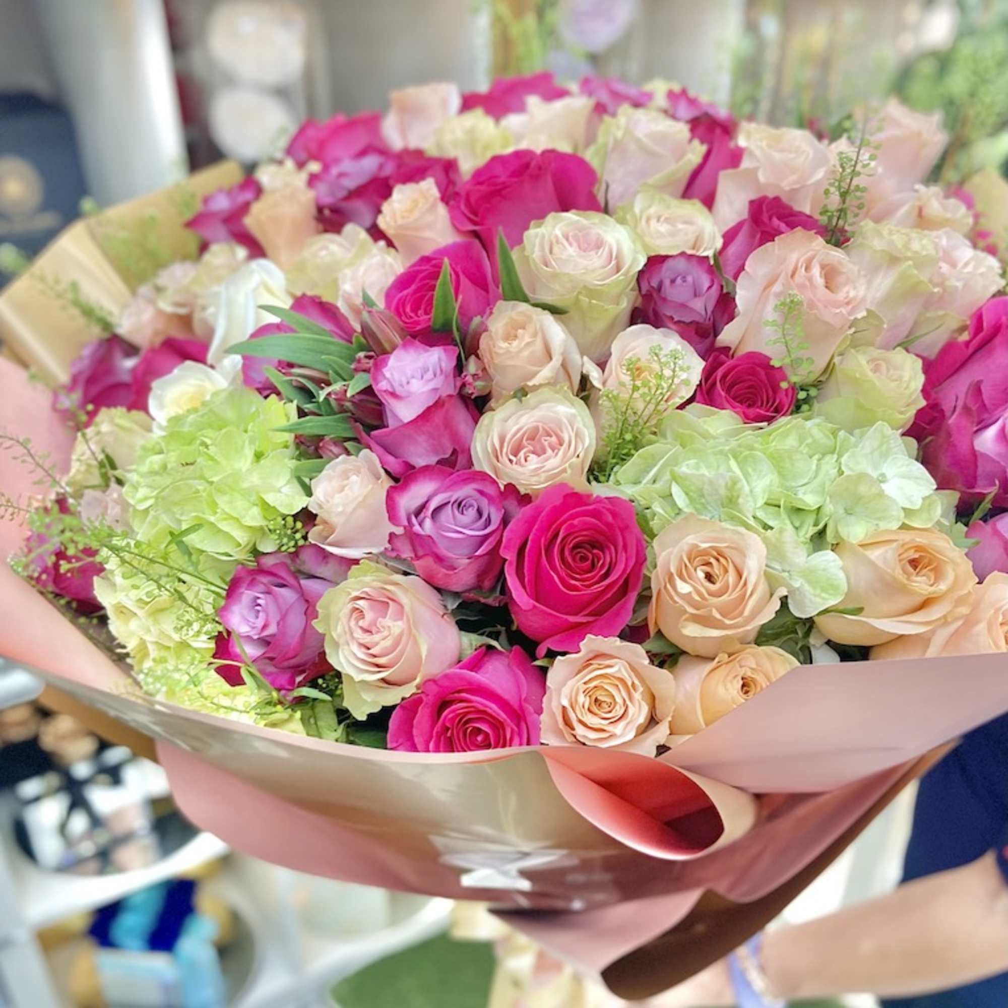 Large hand-tied bouquet of pink, peach, and green roses and hydrangeas wrapped in glossy pink paper.