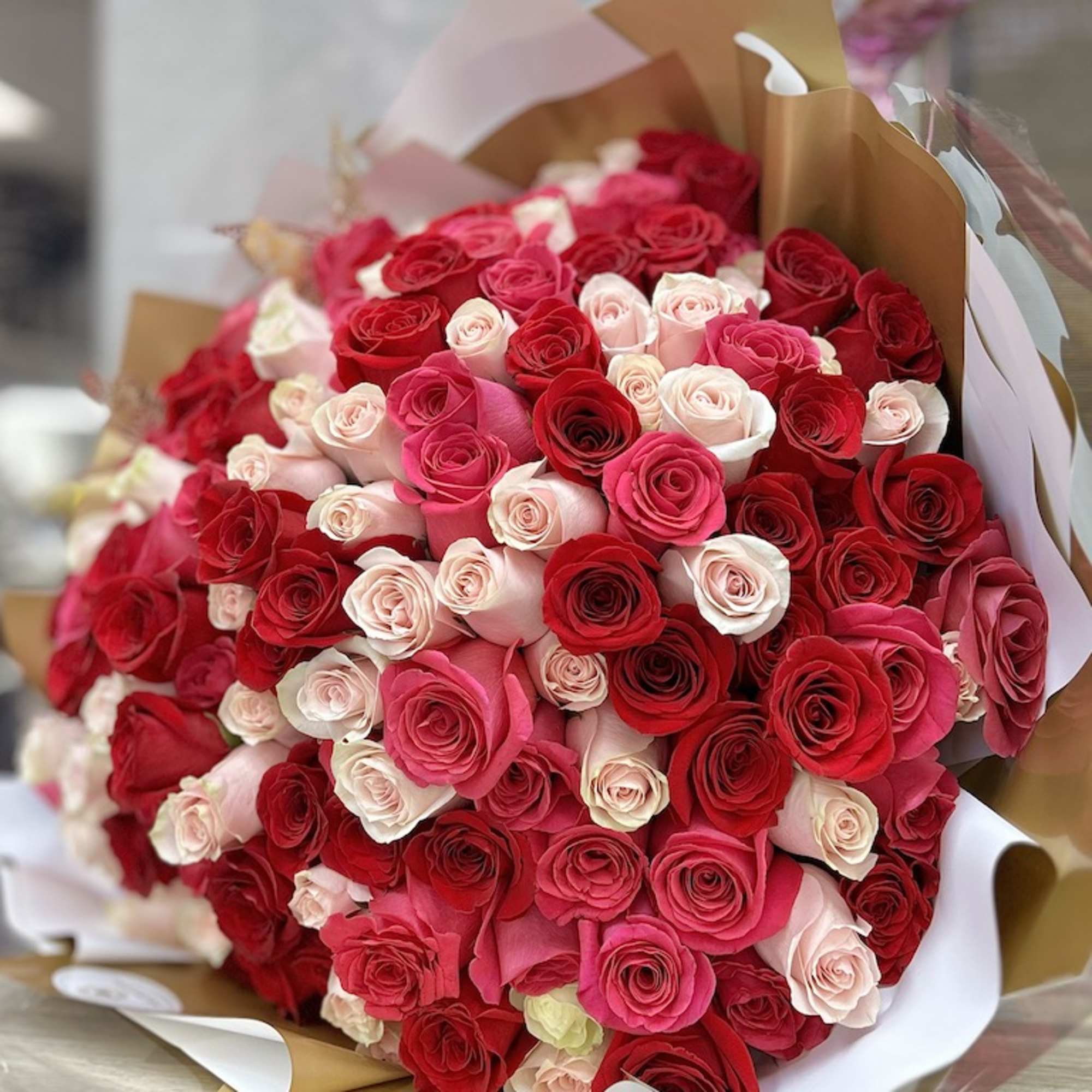 Large round bouquet of red, hot pink, and pale roses wrapped in white and gold paper