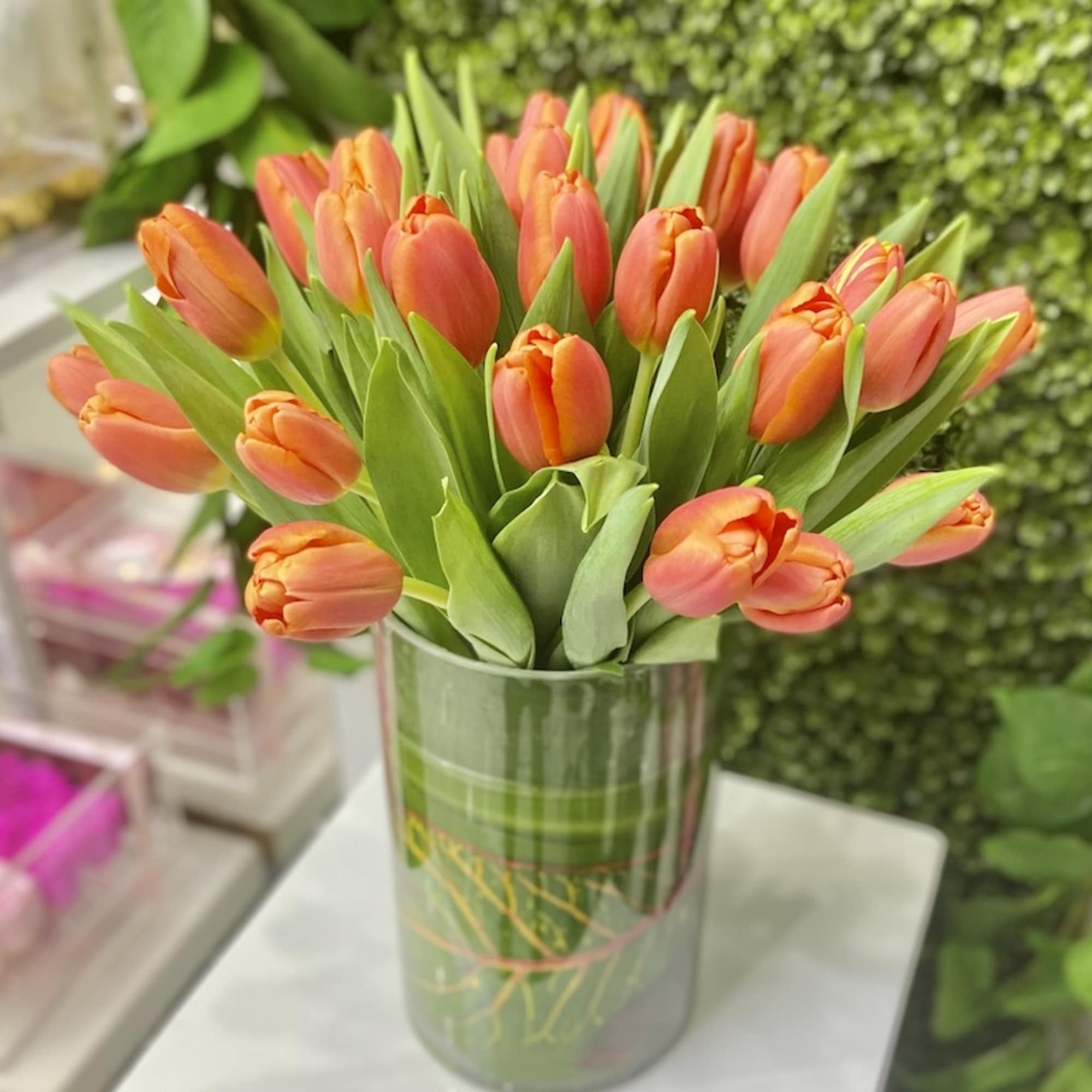 Cluster of orange tulips arranged in a green-wrapped glass cylinder vase on a white pedestal
