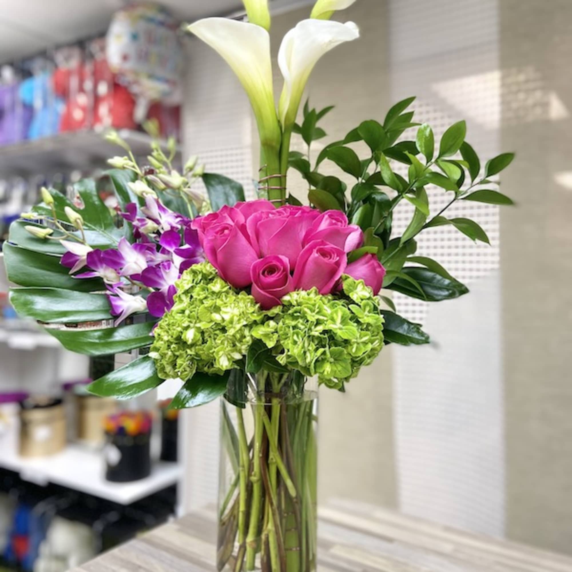 Tall arrangement of white calla lilies, hot pink roses, orchids, and green hydrangeas in a glass vase