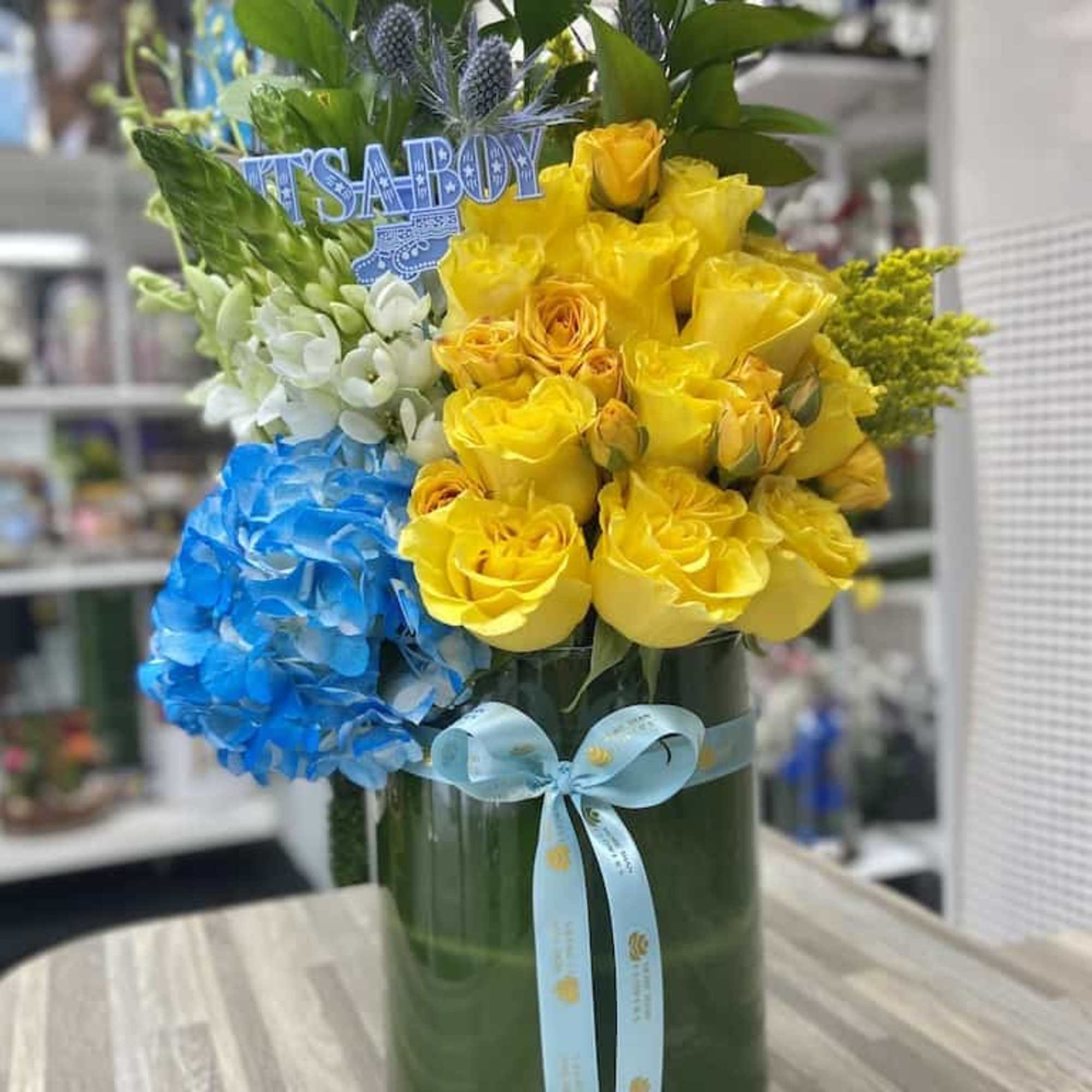 Tall arrangement of yellow roses, blue hydrangea, and white flowers in a green vase with an 'It's a Boy' sign.