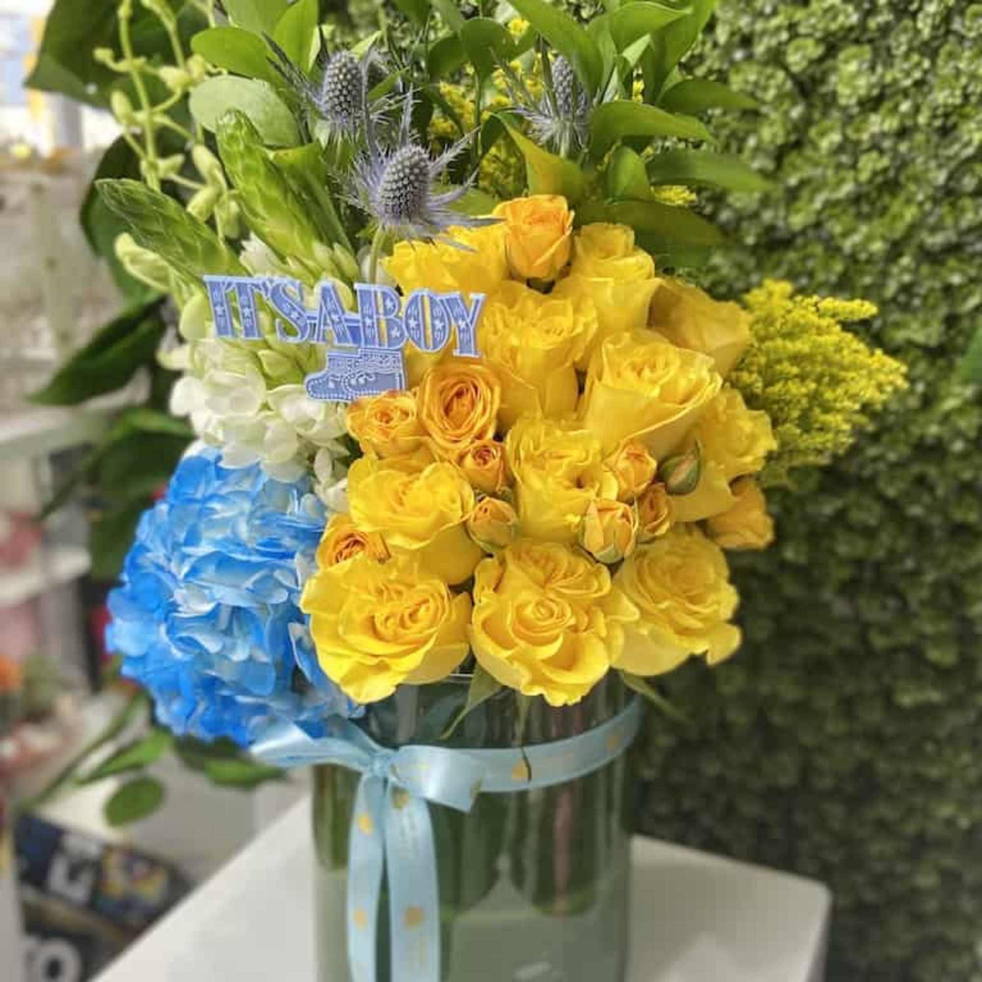 Baby boy themed arrangement with yellow roses and blue hydrangea in a green vase with ribbon