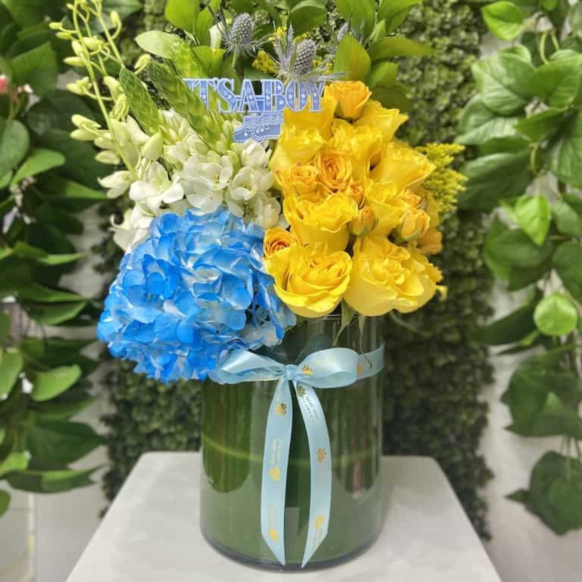 Tall arrangement of yellow roses, blue hydrangea, and white orchids in a glass vase with an 'It’s a Boy' sign.