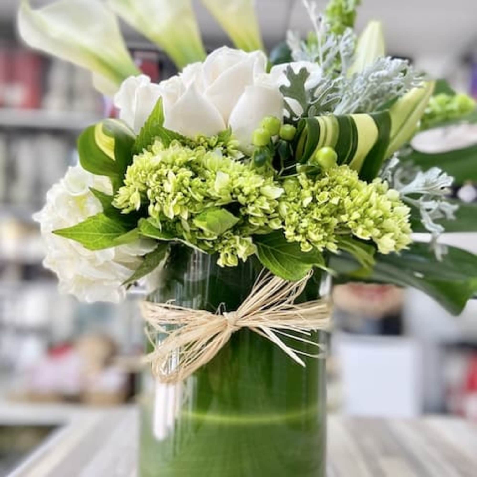 Tall white and green arrangement with calla lilies and roses in a glass vase wrapped with green leaves