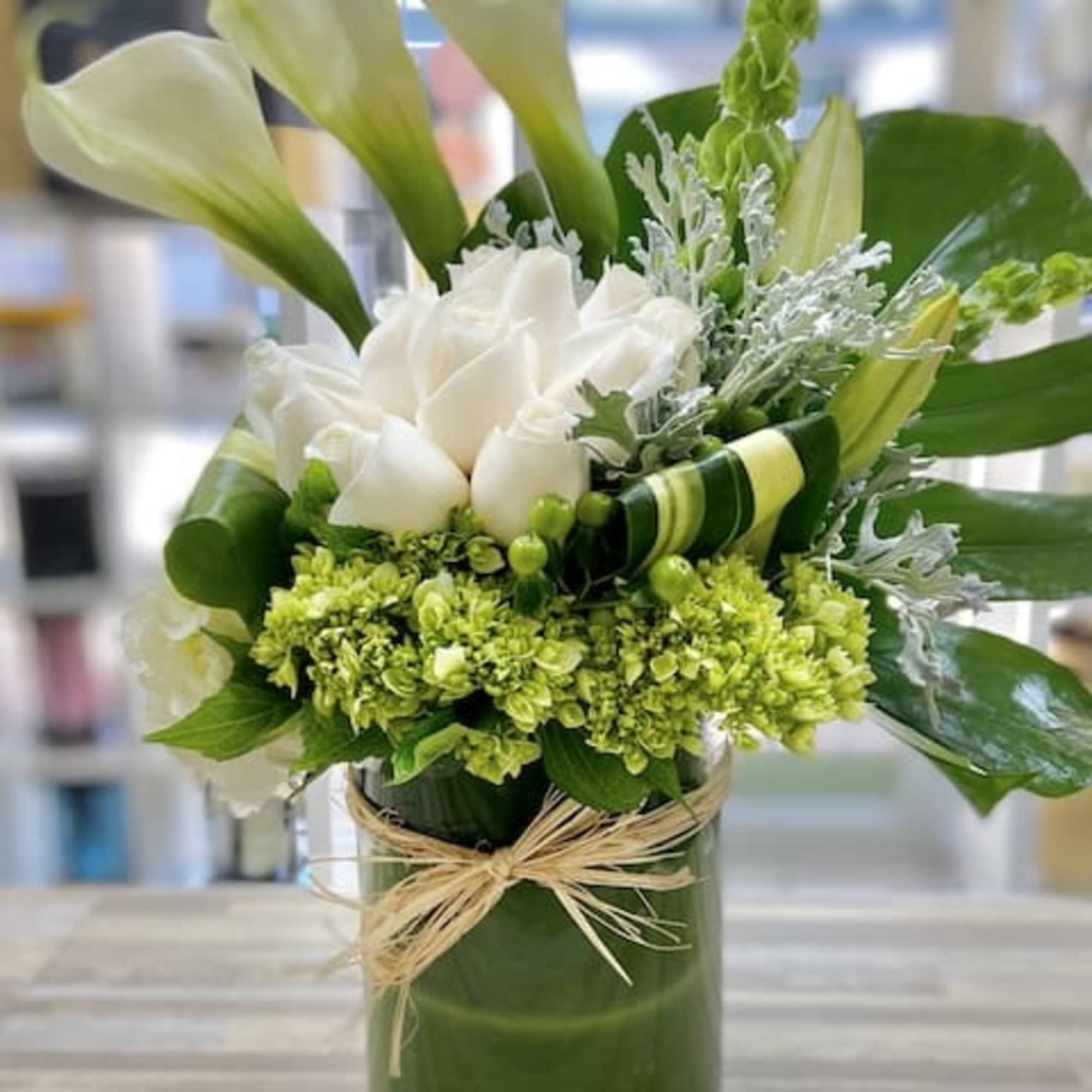 White roses and green blooms with calla lilies in a green-lined glass cylinder vase tied with raffia