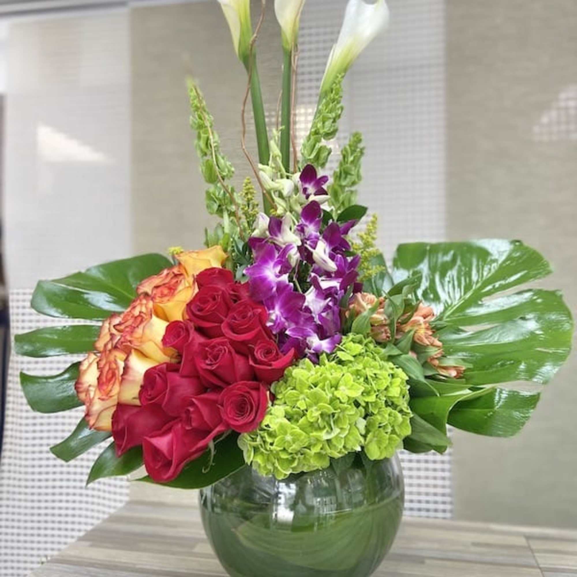 Tall arrangement with white calla lilies, red and orange roses, purple orchids and green hydrangea in a glass vase