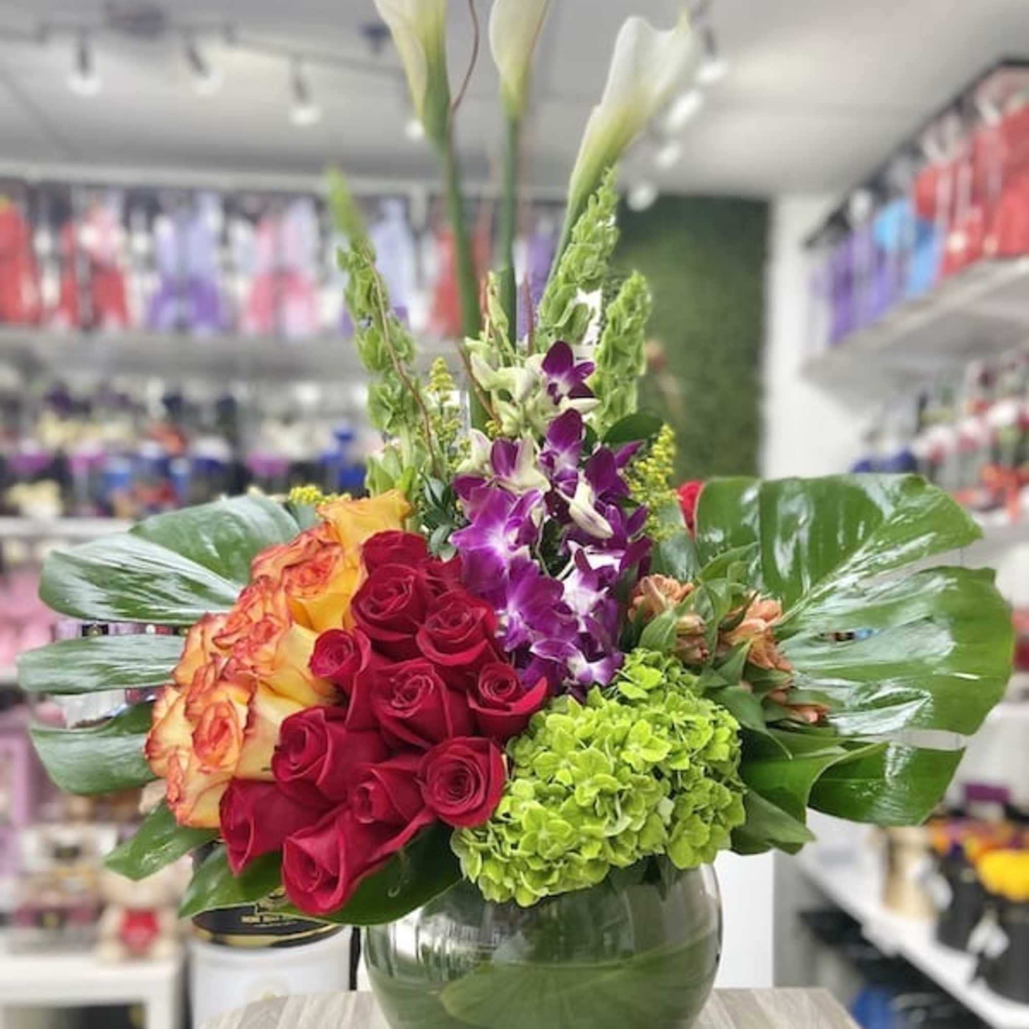 Tall mixed arrangement with red roses, white calla lilies, orchids, hydrangea and large glossy leaves in a round glass vase