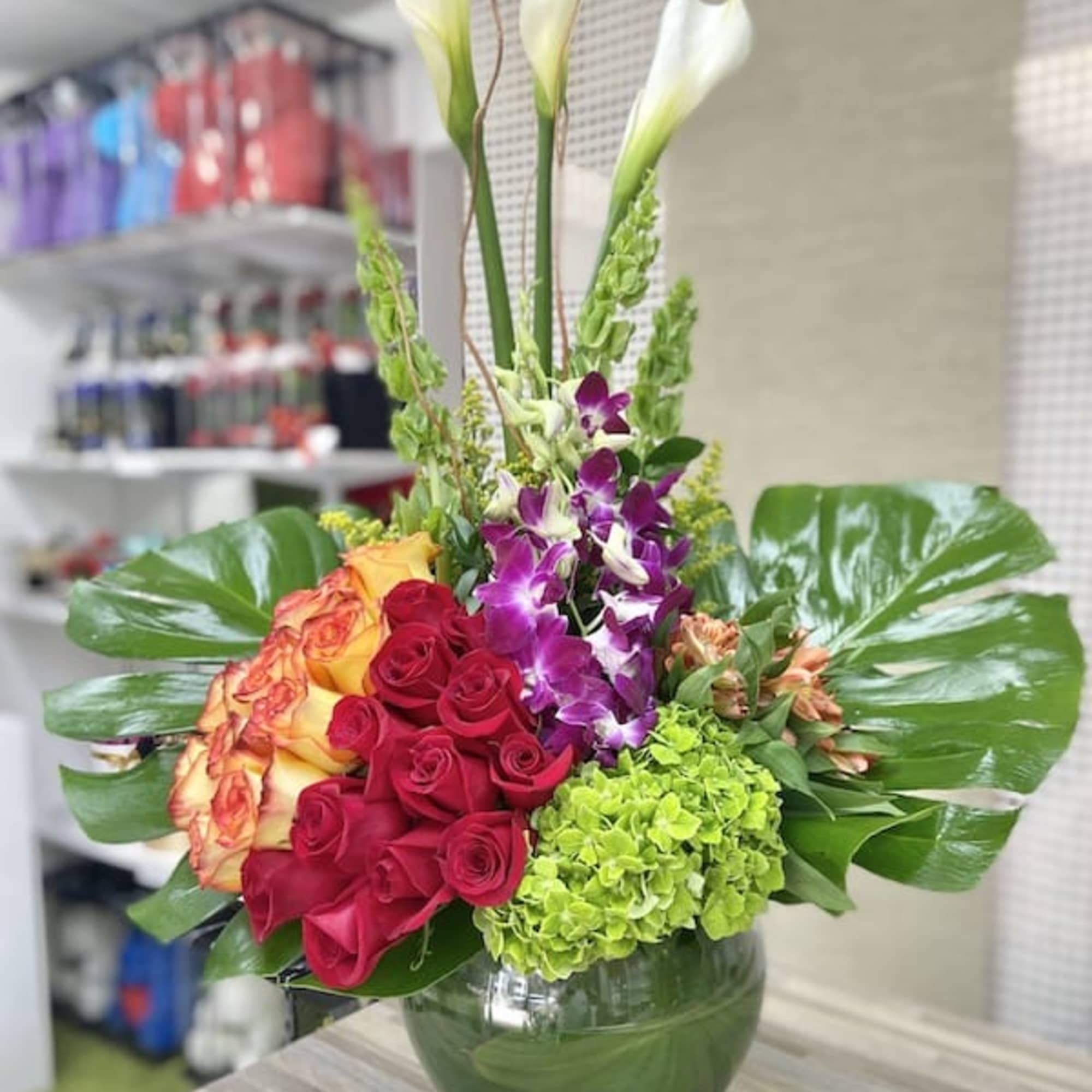 Tall arrangement with white calla lilies, red and orange roses, orchids and green hydrangea in a round glass vase