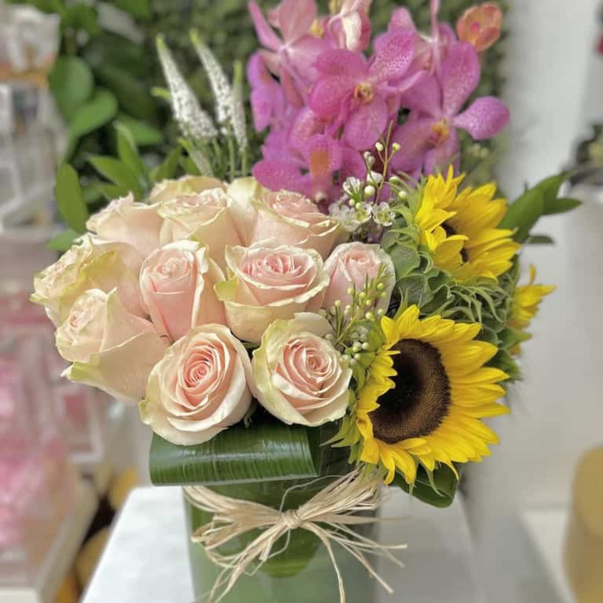 Compact arrangement of blush roses, yellow sunflowers and pink orchids in a green-wrapped glass vase with raffia bow