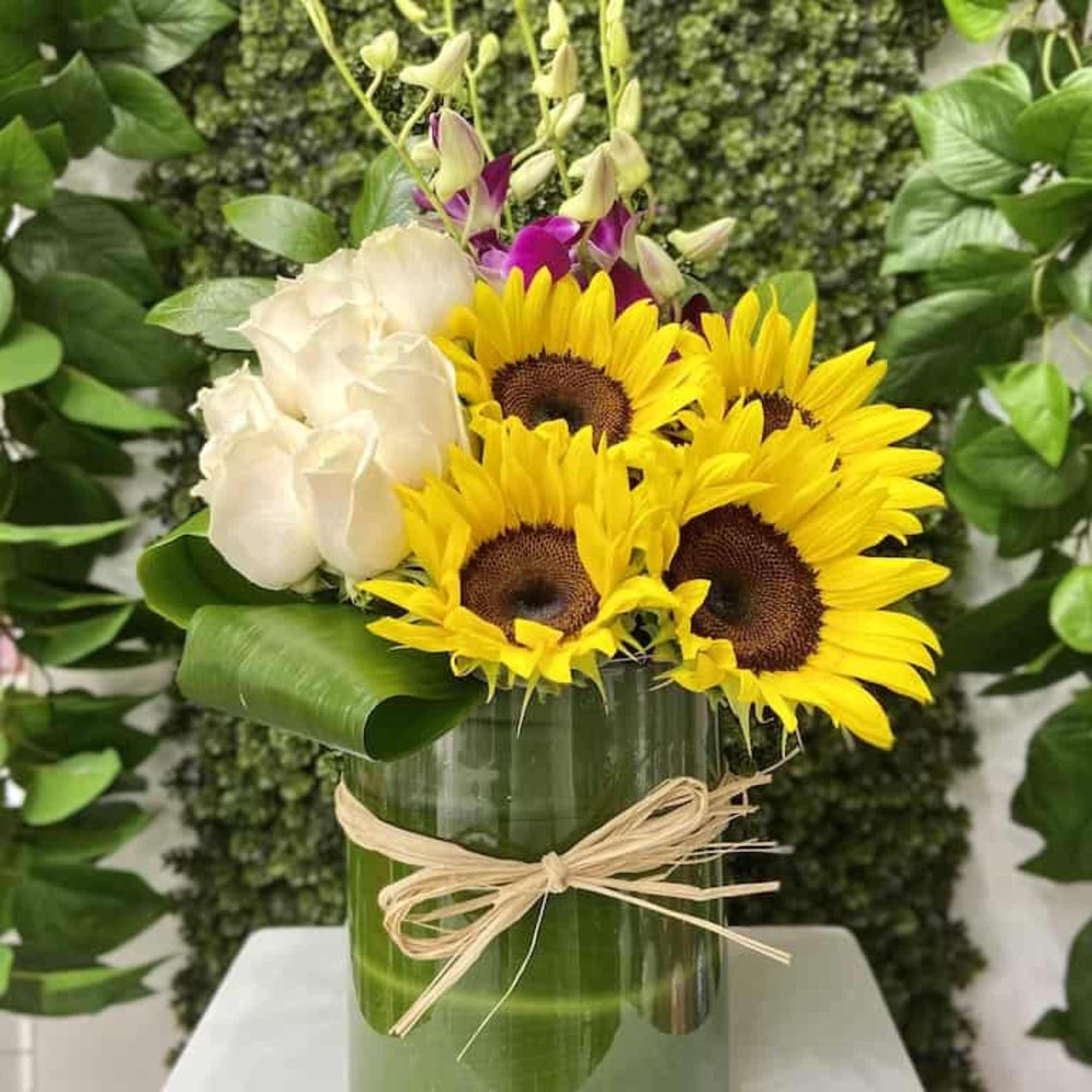 Arrangement of sunflowers, white roses and orchids in a glass vase wrapped with a green leaf and raffia bow