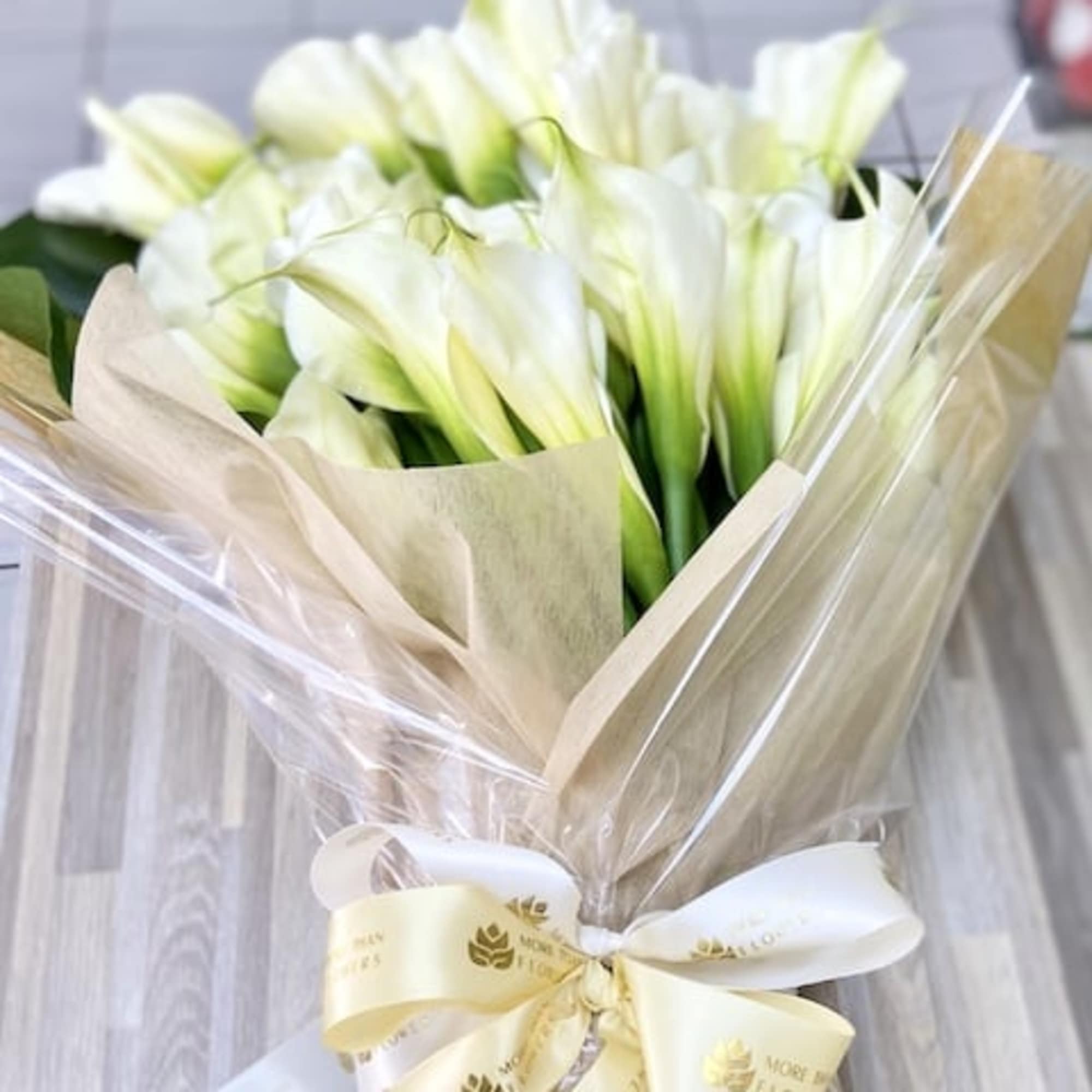 Bouquet of white calla lilies wrapped in kraft paper and clear cellophane with cream ribbons.