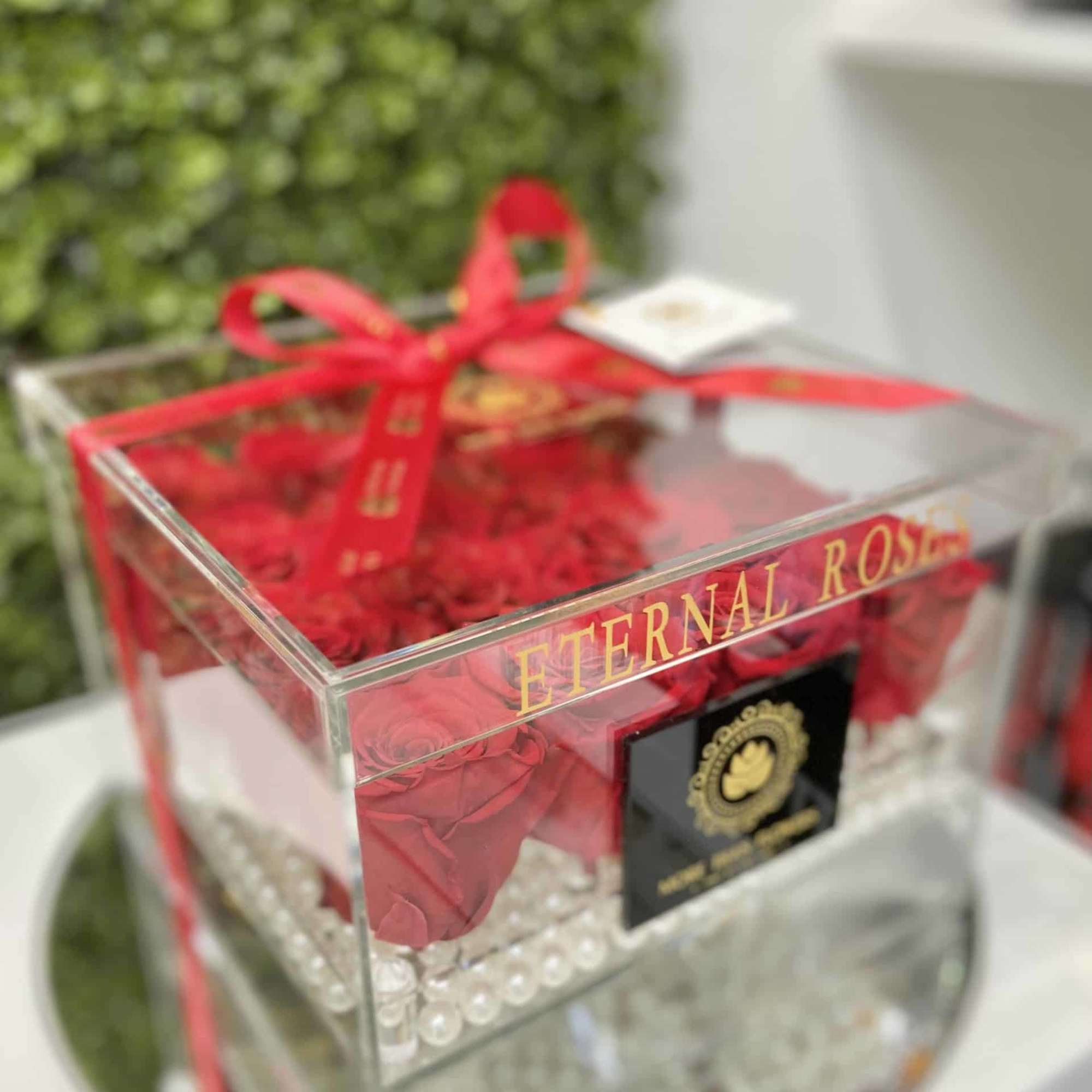 Clear acrylic box of red roses with pearl accents and red ribbon labeled Eternal Rose