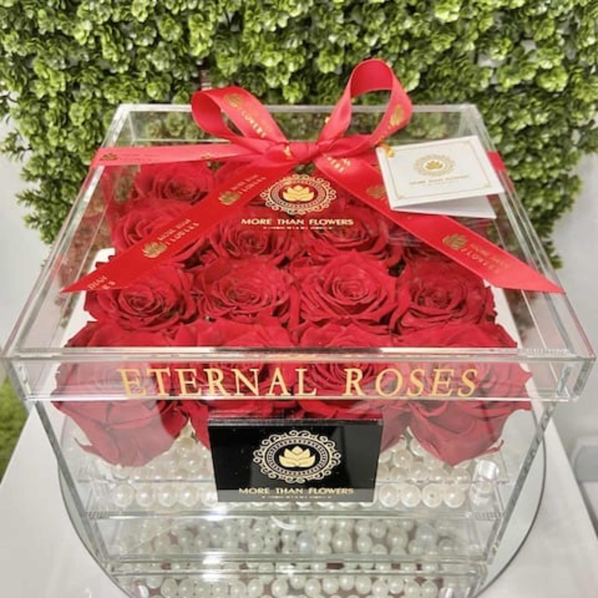 Clear acrylic box of red roses with pearls and a red ribbon labeled Eternal Roses