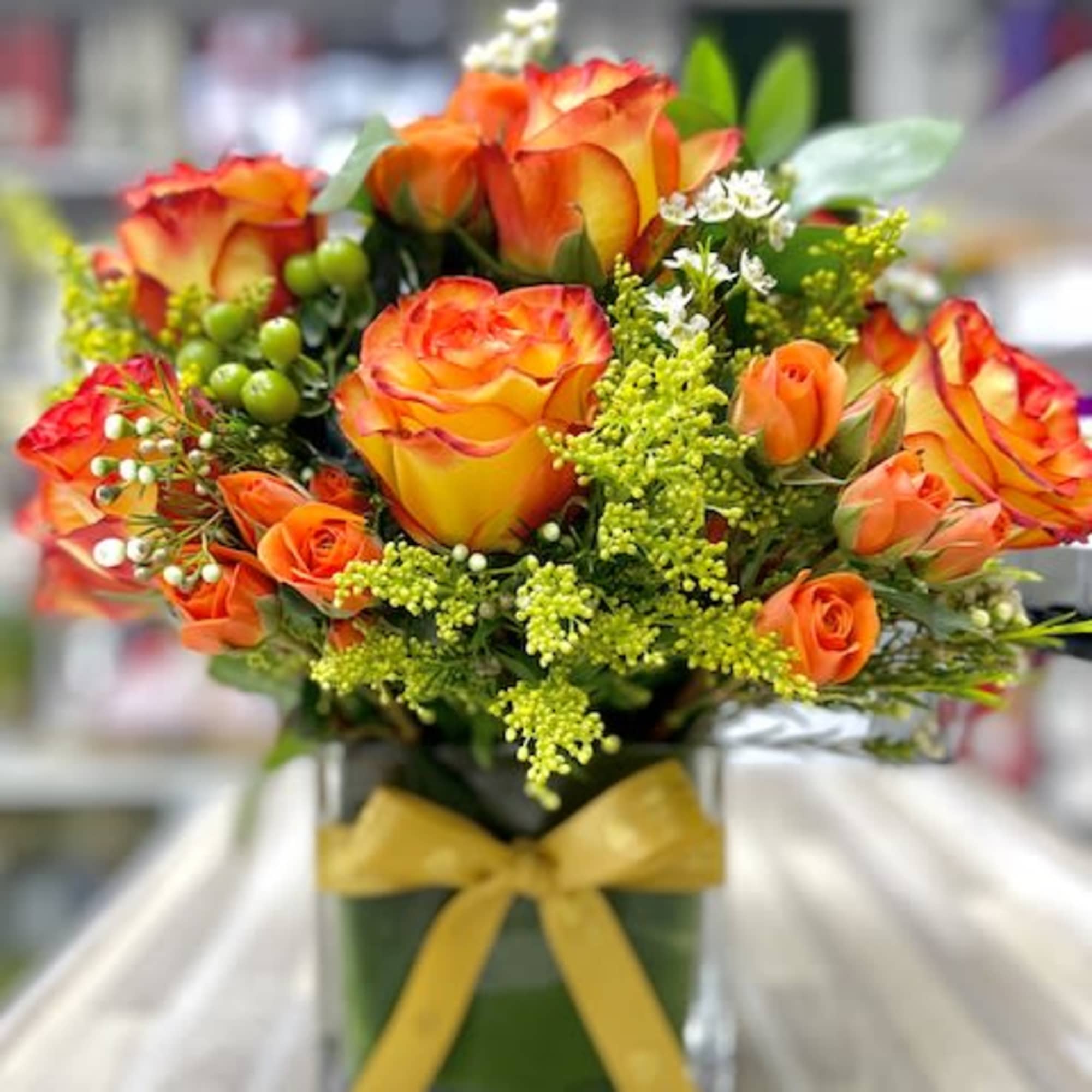 Orange and yellow roses with filler flowers in a glass cube vase tied with a yellow bow.