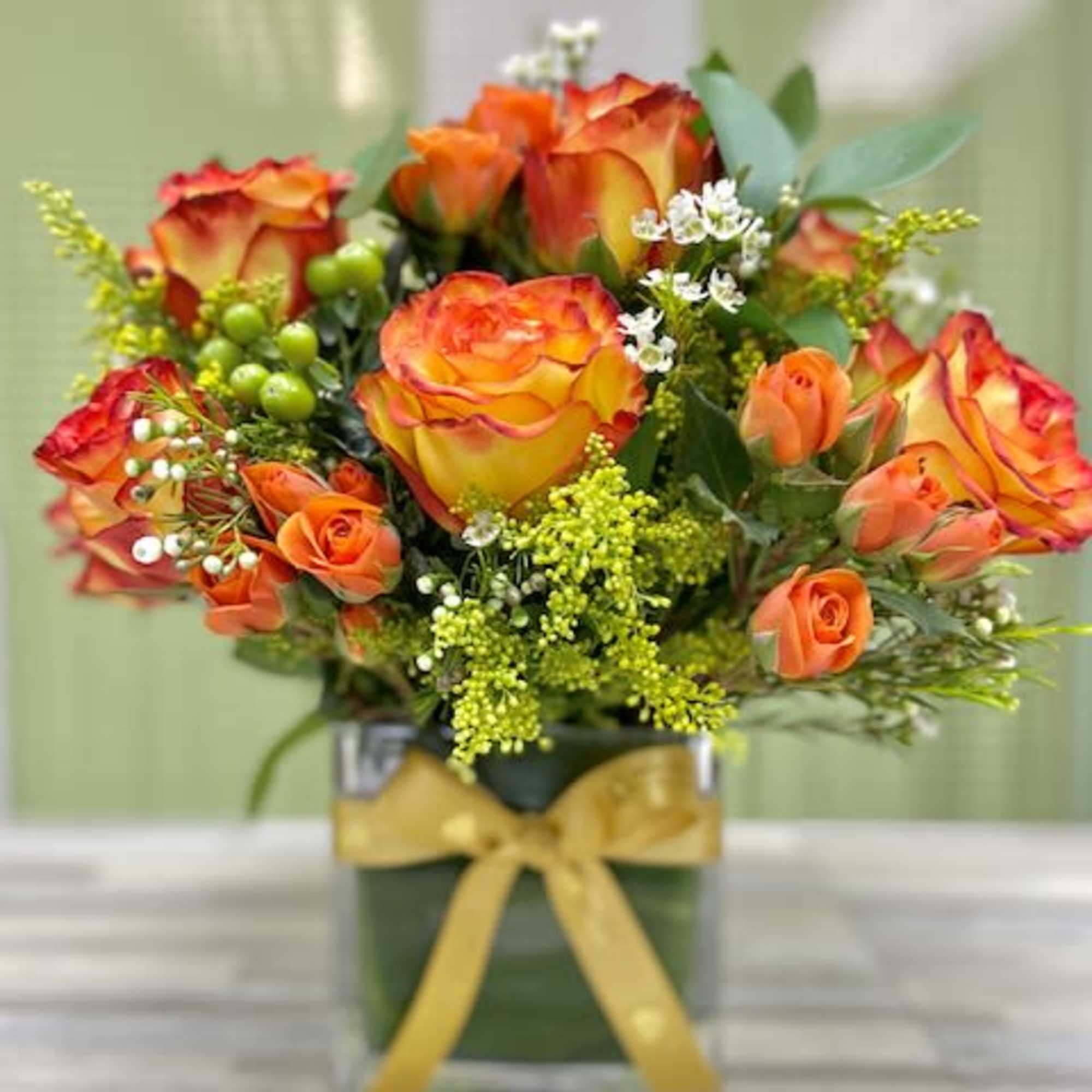 Compact arrangement of orange and yellow roses with white accents in a glass cube vase with yellow ribbon