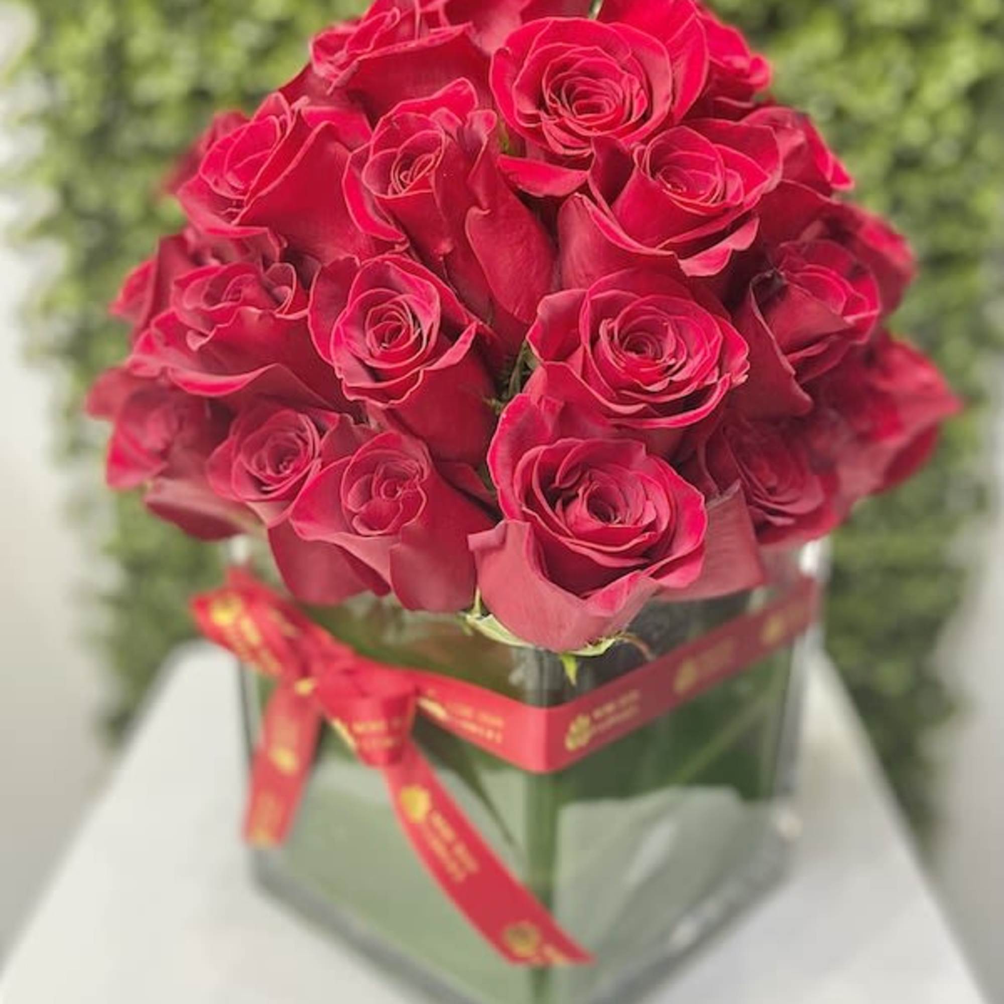 Dense bouquet of red roses arranged in a square glass vase wrapped with a red ribbon.