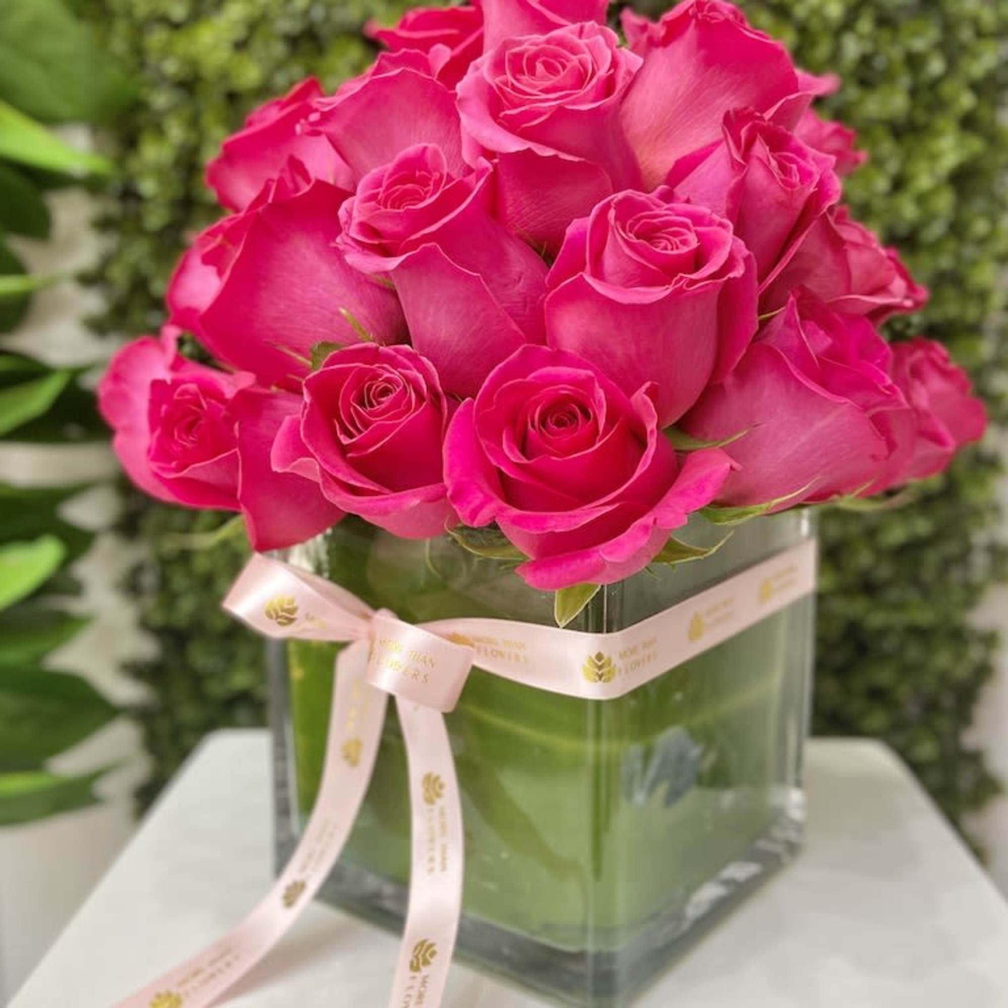 Compact arrangement of bright pink roses in a square glass vase with a pale pink ribbon bow.