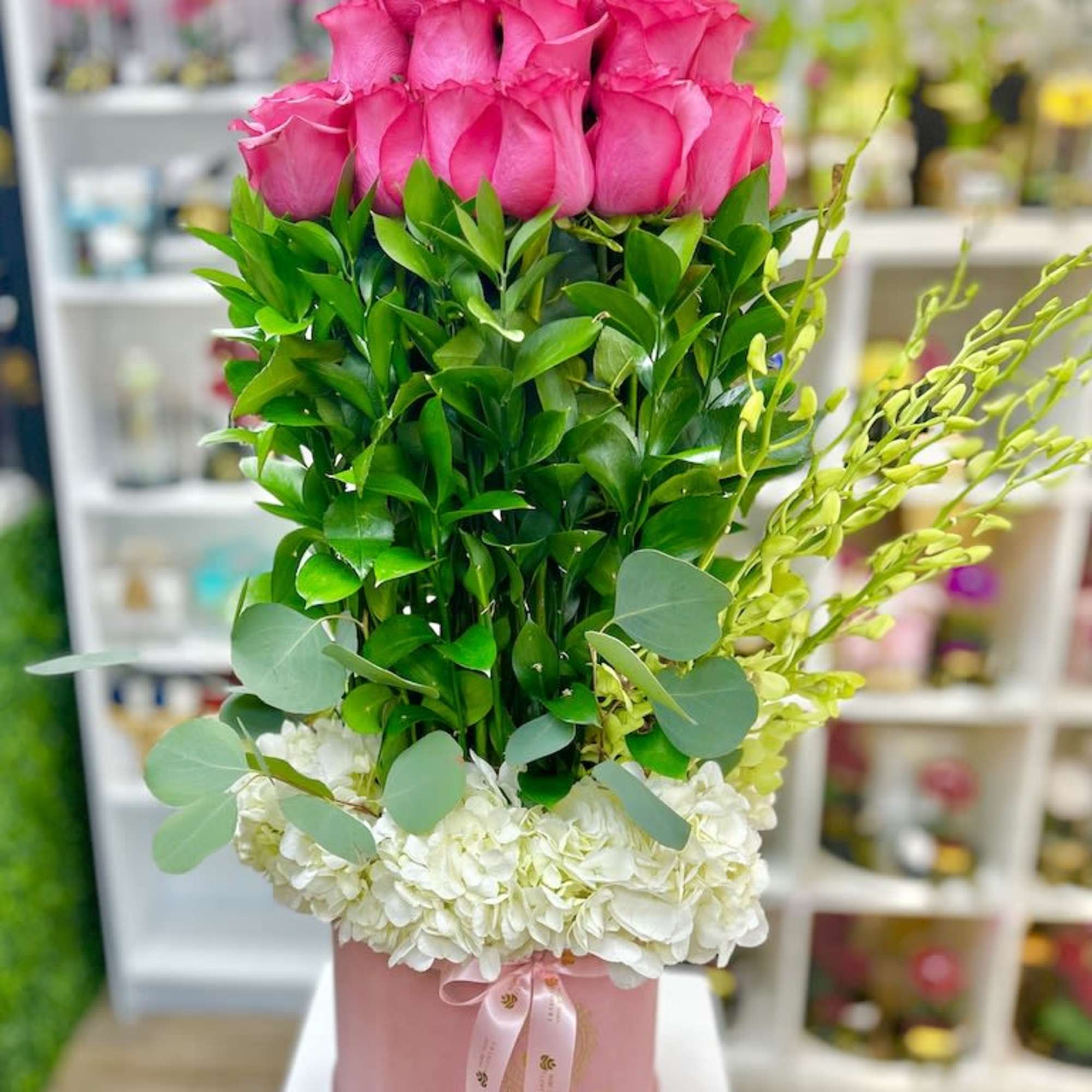 Tall arrangement of pink roses, layered greenery, and white hydrangeas in a pink hatbox.