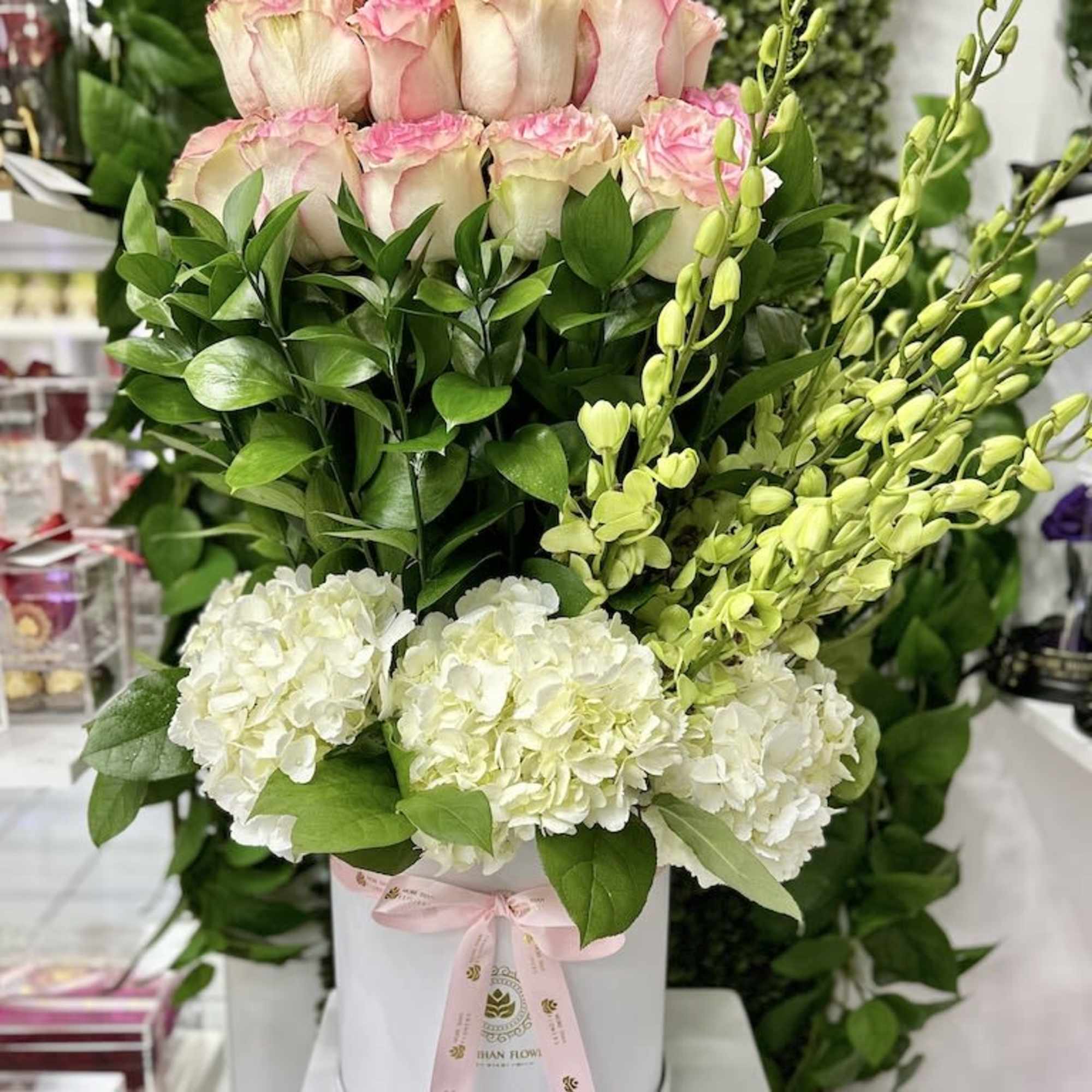 Tall arrangement of pink-tipped roses, white hydrangeas, and green orchids in a white hatbox with pink ribbon