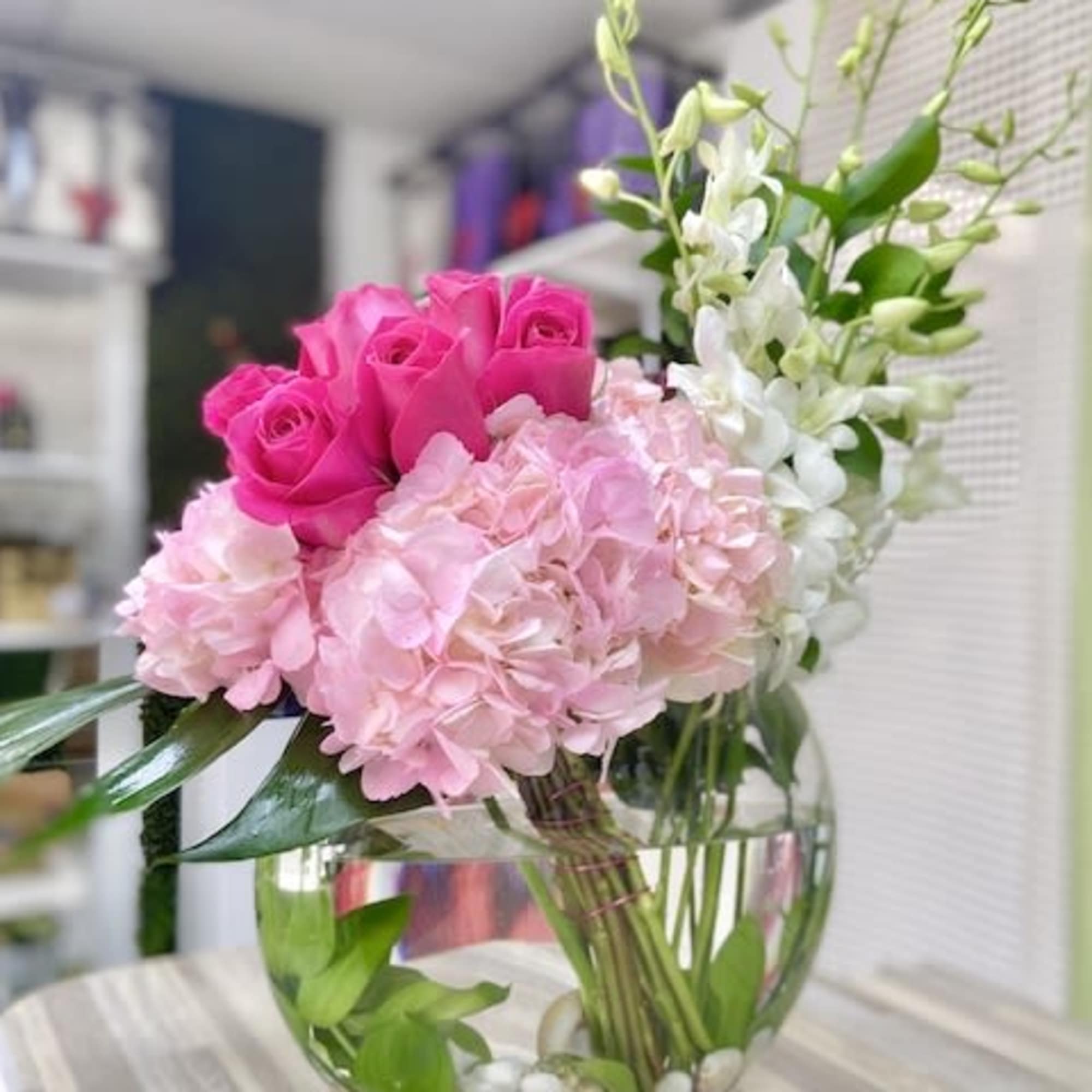 Round glass vase with pink roses, pink hydrangeas, and white orchids arranged low with visible stems and stones.