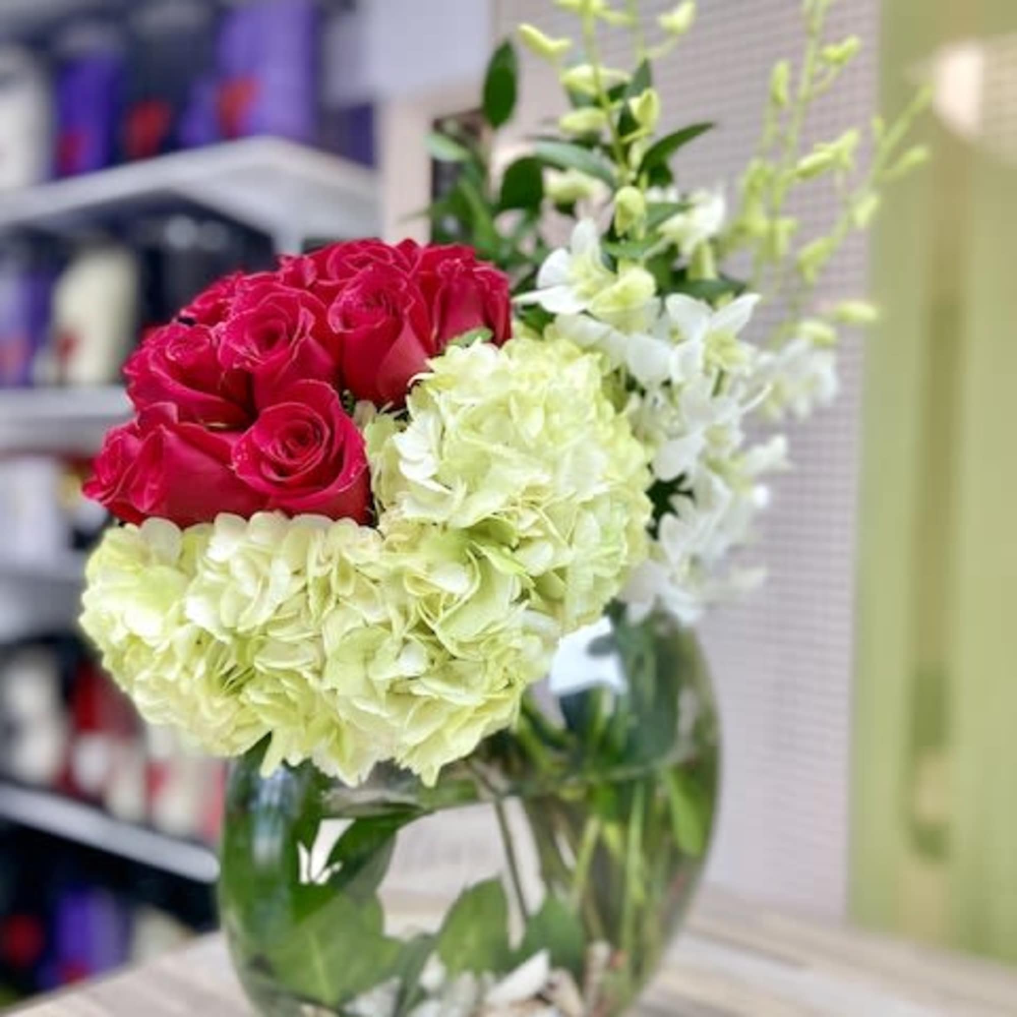 Asymmetrical arrangement of red roses, green hydrangeas, and white orchids in a round glass vase with shells.