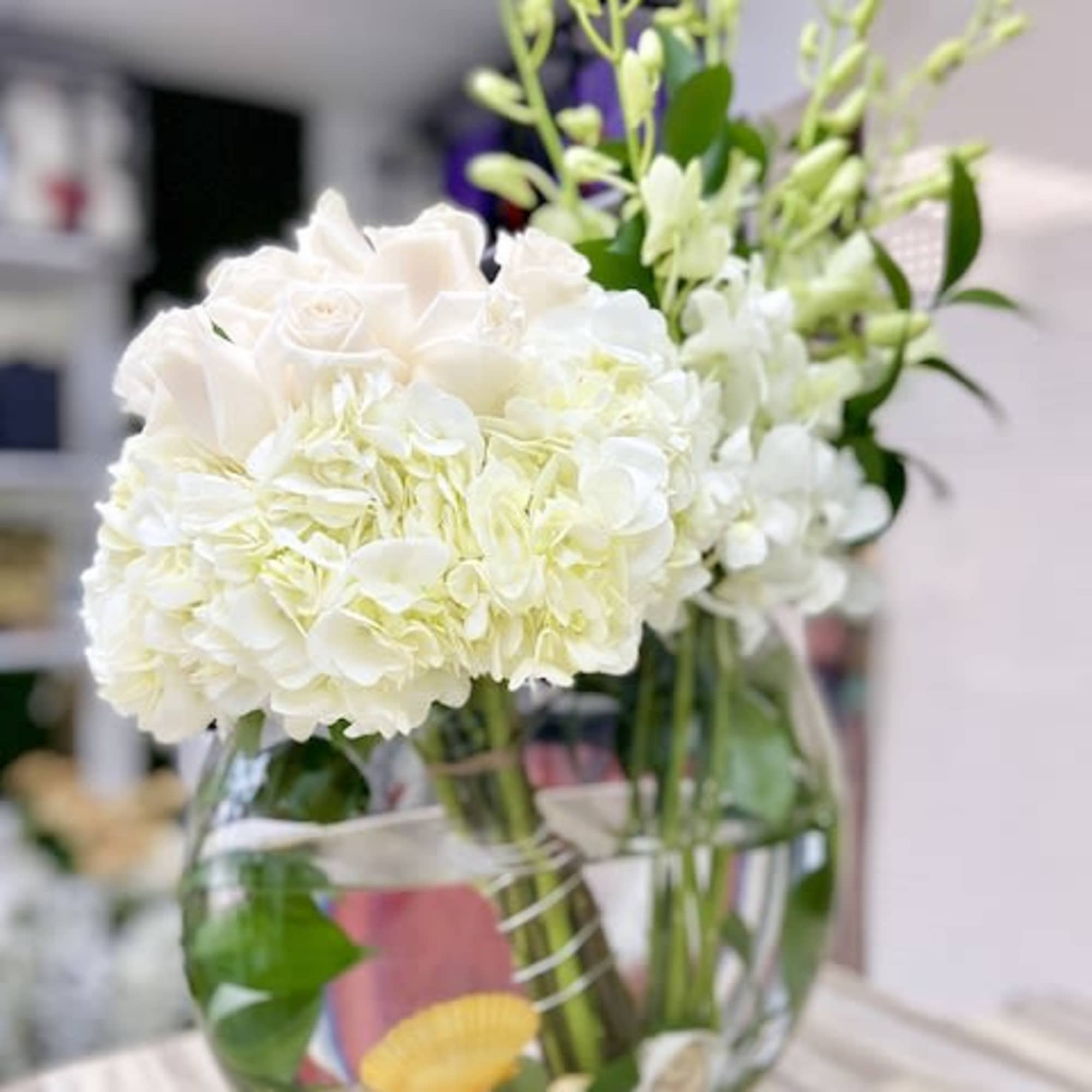 Round glass vase of white hydrangeas, roses, and orchids with seashells in the water at the base.