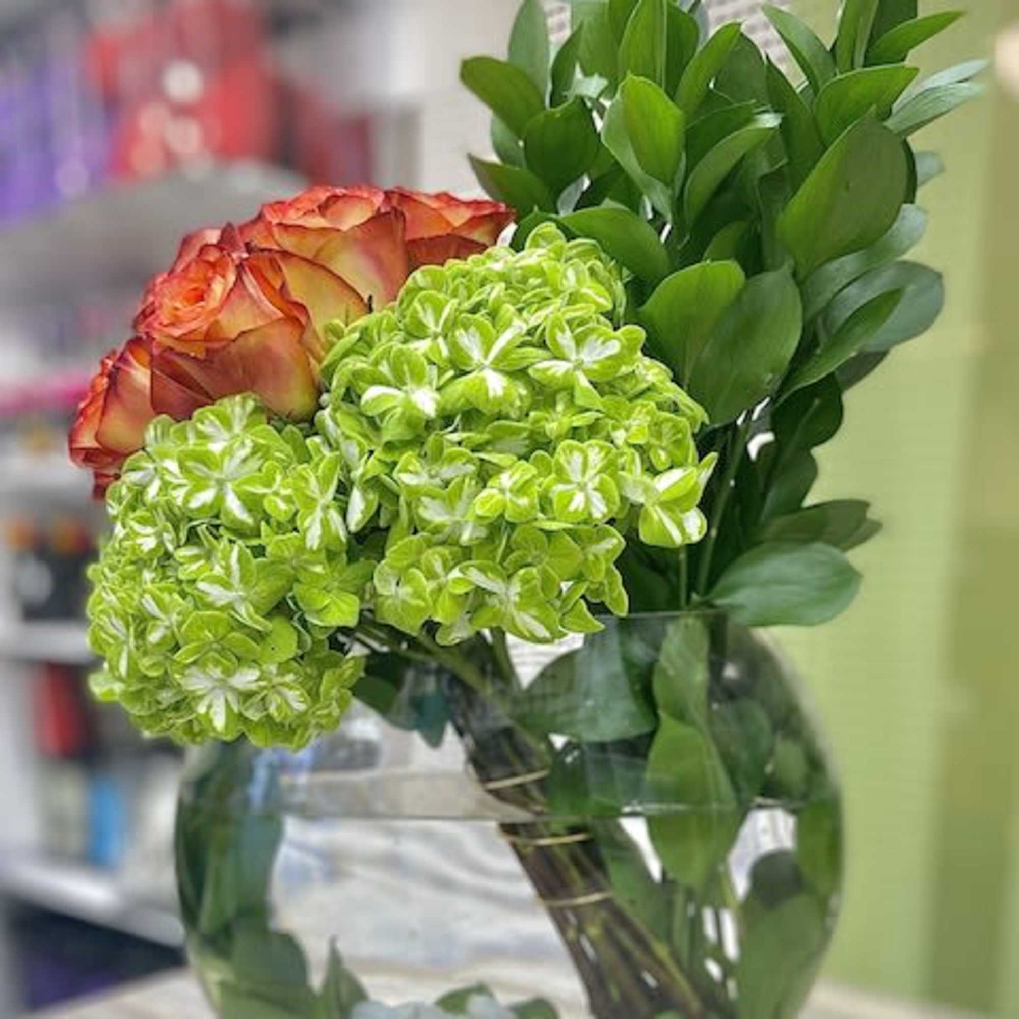 Green hydrangeas and orange roses with tall foliage arranged in a round clear glass vase